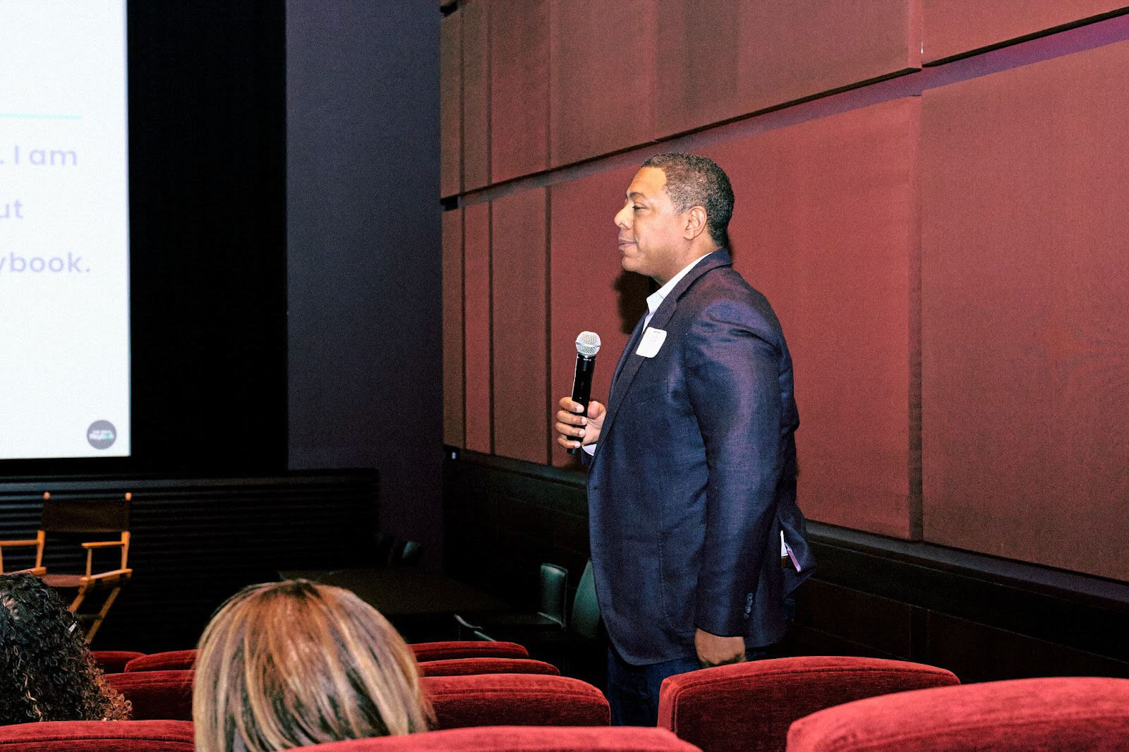 A man in formal attire standing in a theater-style room, holding a microphone, and addressing the audience during an event or discussion.