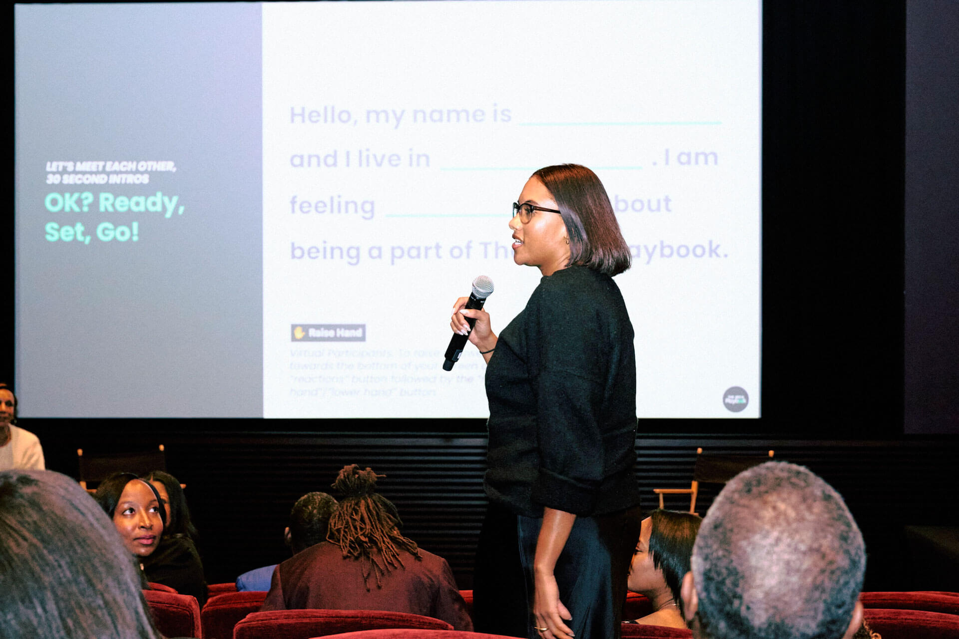A presenter stands before a projected slide introducing an interactive session, holding a microphone while speaking to the seated audience.