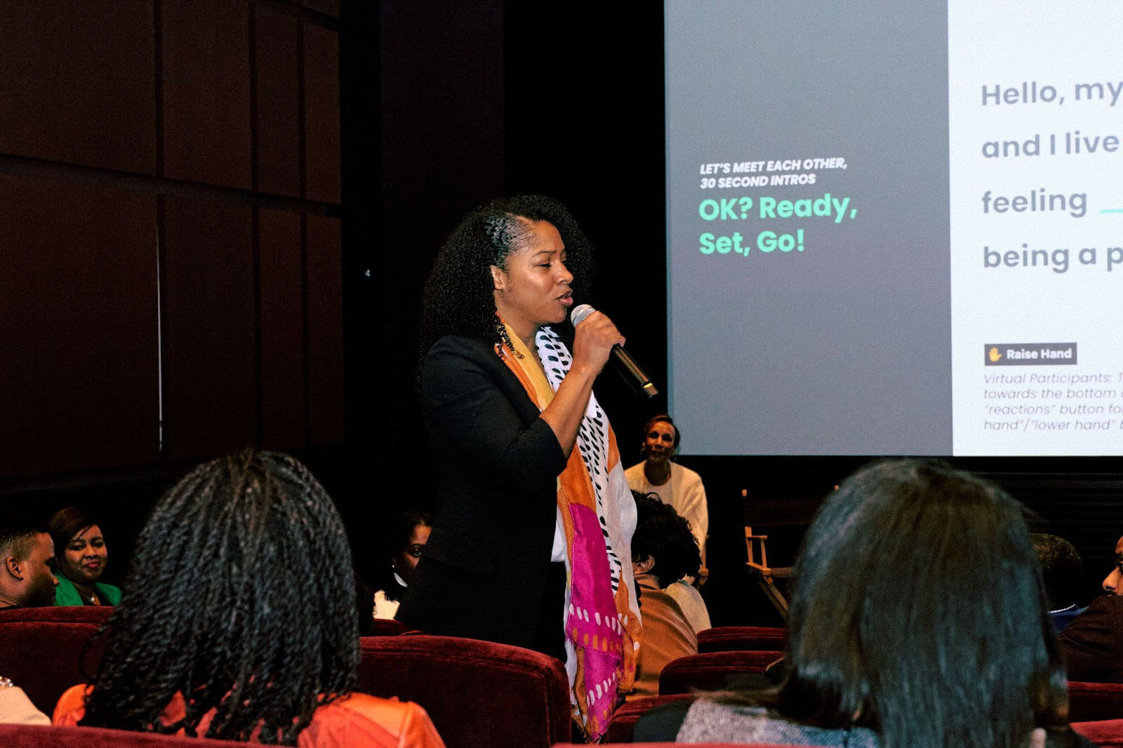 A woman in a colorful scarf and black blazer engages the audience while speaking into a microphone.