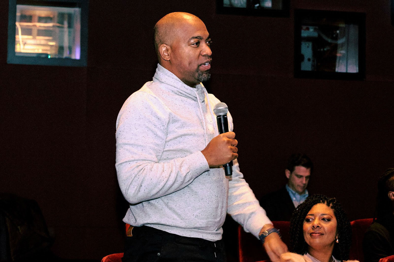 A man in a gray sweater stands with a microphone in hand, actively addressing the audience during a session.