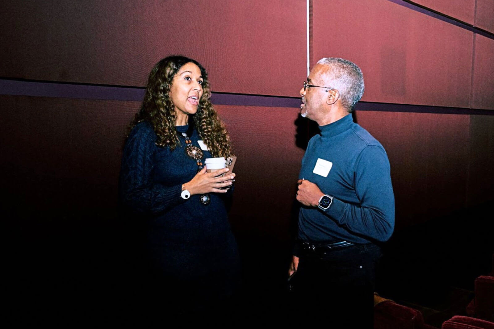 Two individuals engaged in a friendly conversation during a professional event, one holding a cup of coffee.
