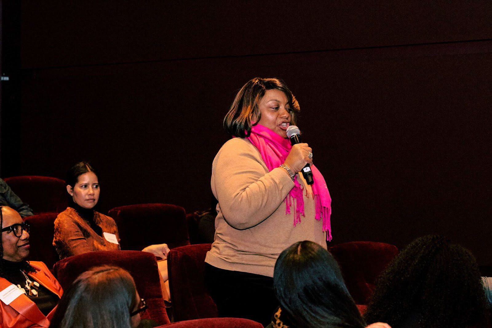 A woman in a pink scarf speaking into a microphone while addressing a seated audience in a theater setting.