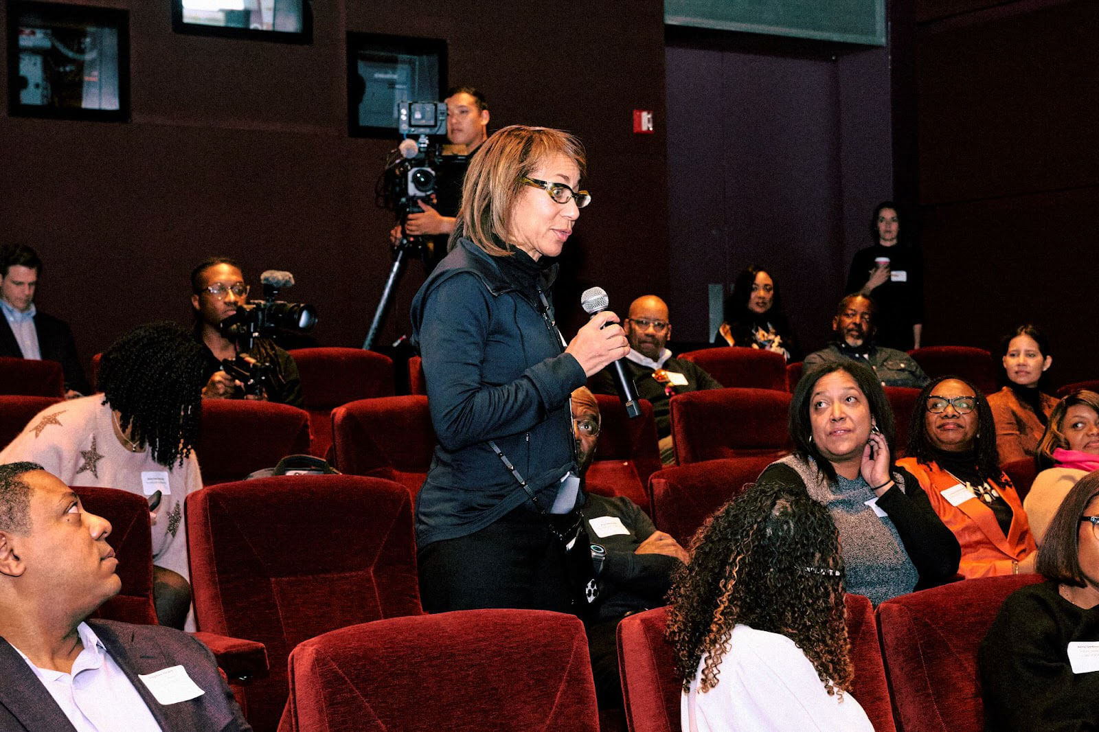 A woman speaking into a microphone with cameras and attendees visible in the background of a theater-style setting.