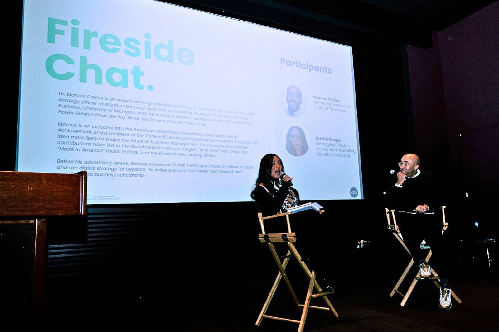 A speaker on stage addressing an audience during a fireside chat, with a large screen behind displaying participant details.