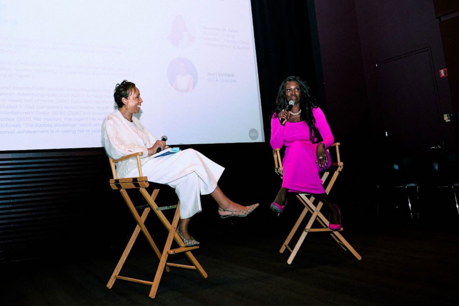 Two women on stage in a conversation, one in a white outfit and the other in a bright pink dress, sharing insights during a panel discussion.
