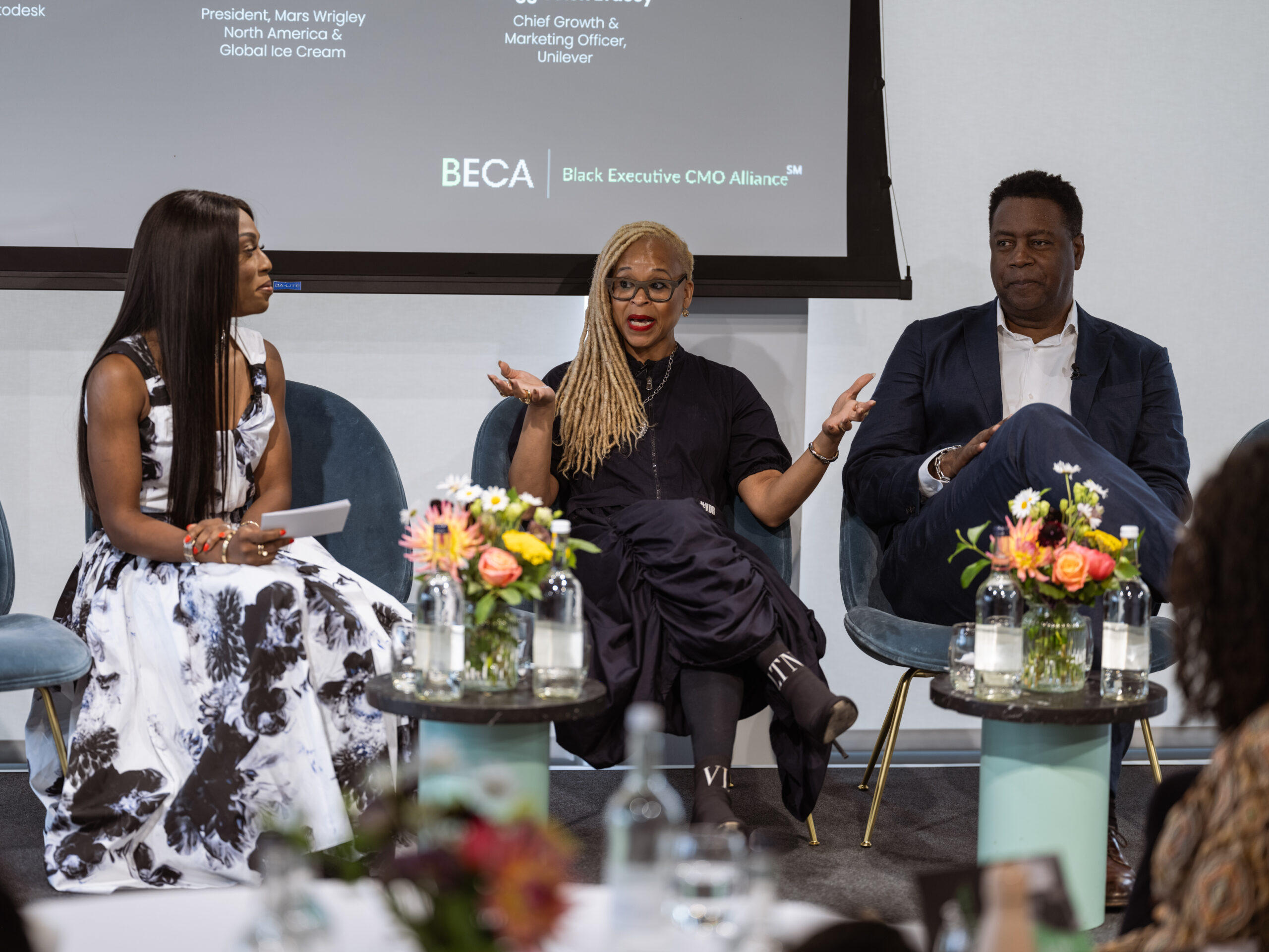 Three panelists seated on stage in front of a screen displaying “BECA | Black Executive CMO Alliance.” The woman in the center, wearing all black and gesturing with her hands, is flanked by a woman in a black-and-white dress on the left and a man in a dark suit on the right. Colorful flowers and water bottles rest on small tables in front of them.