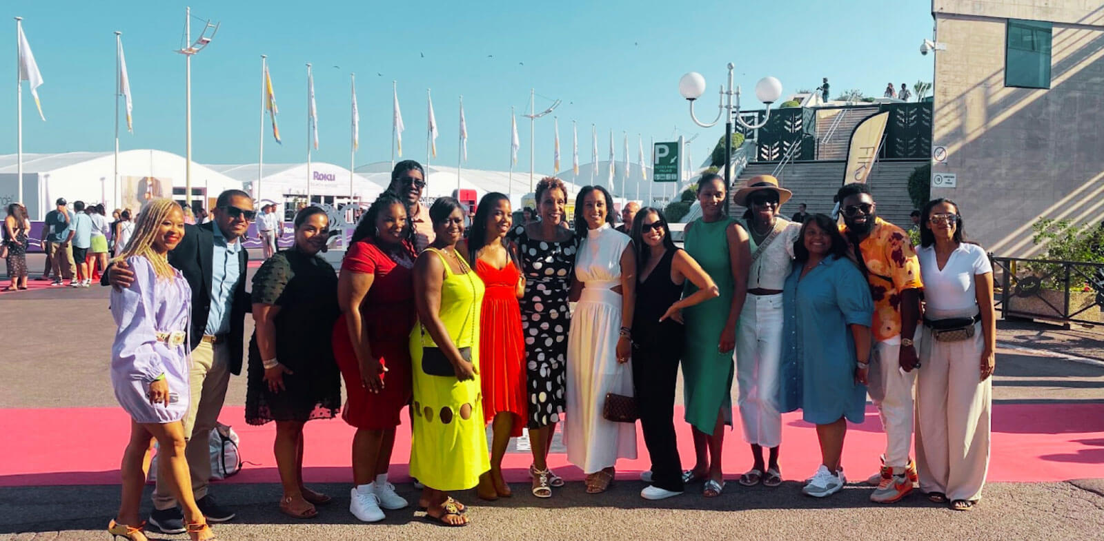 A group of individuals posing together on a red carpet, with flags and event tents visible in the background under a sunny sky.