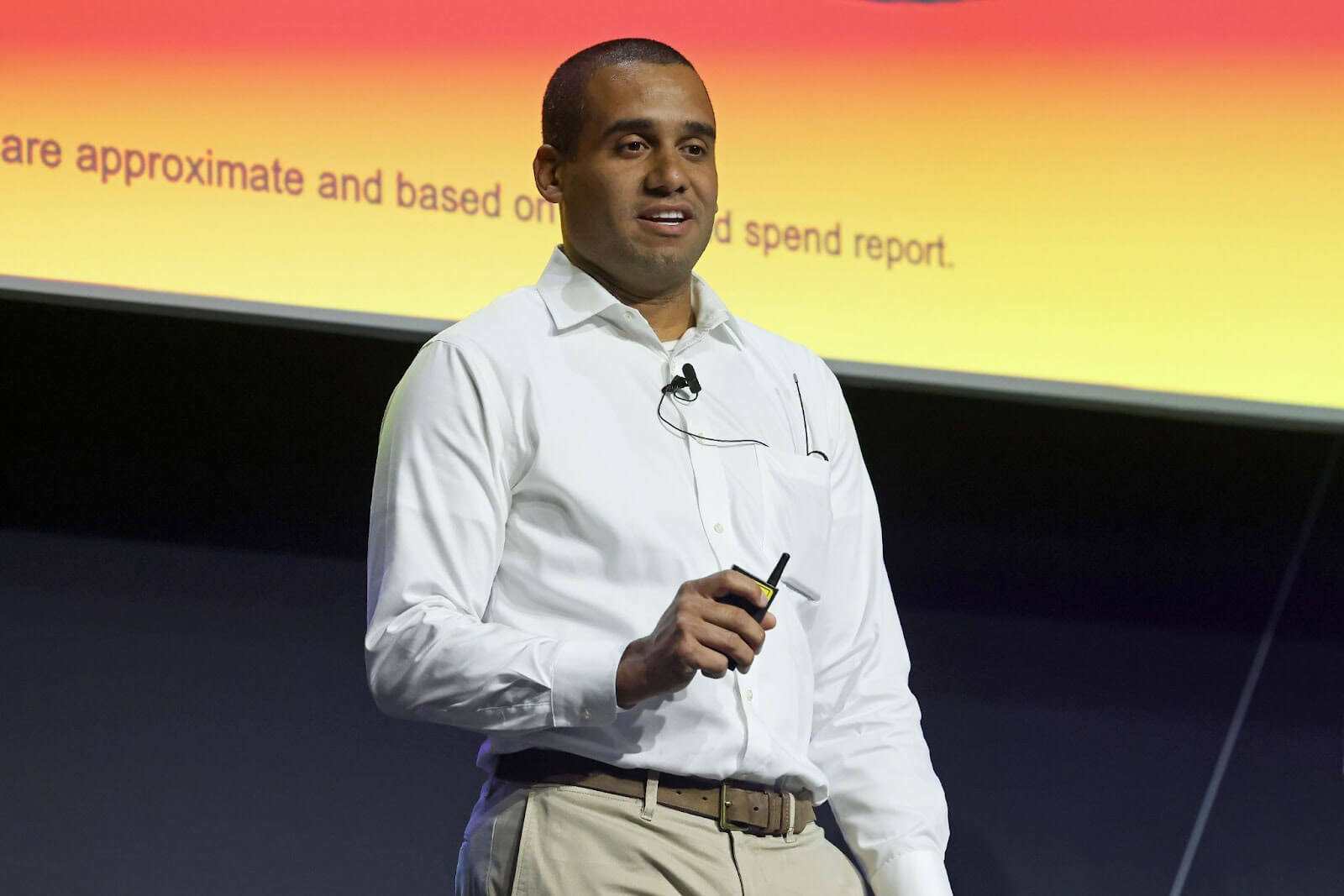 A man in a white shirt and khakis giving a presentation on stage, holding a clicker with a brightly colored slide in the background.