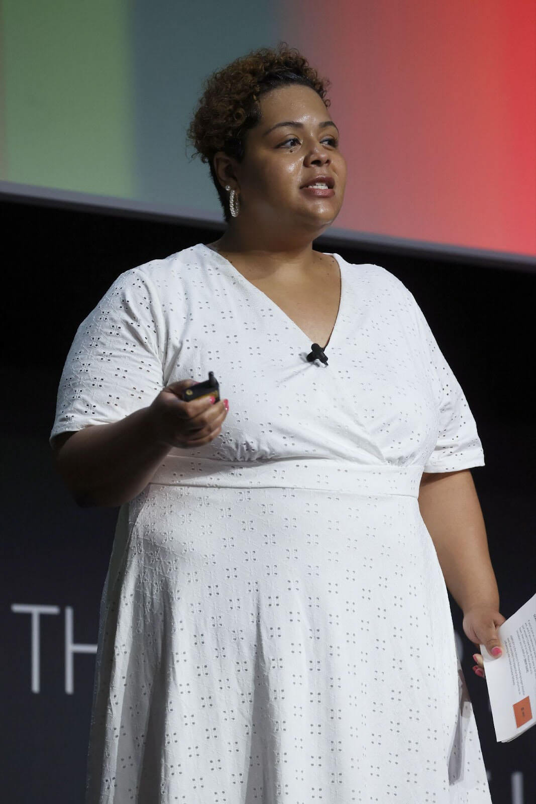 A woman in a white dress giving a presentation on stage, holding a clicker and a paper.