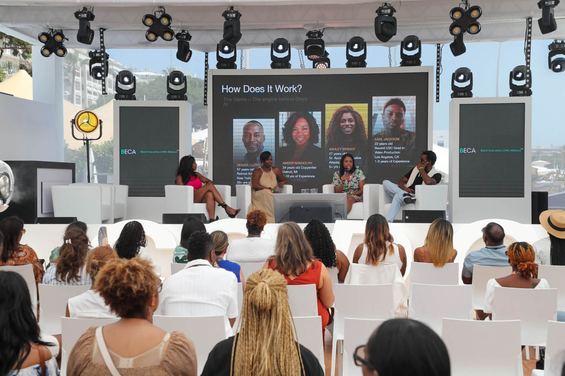 A live panel discussion with four speakers on stage, featuring a large screen displaying profiles and an attentive audience seated in the foreground.