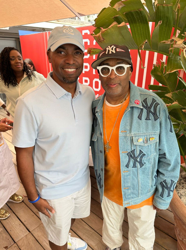 Two individuals, including Spike Lee in a denim jacket and signature white glasses, posing for a photo outdoors.