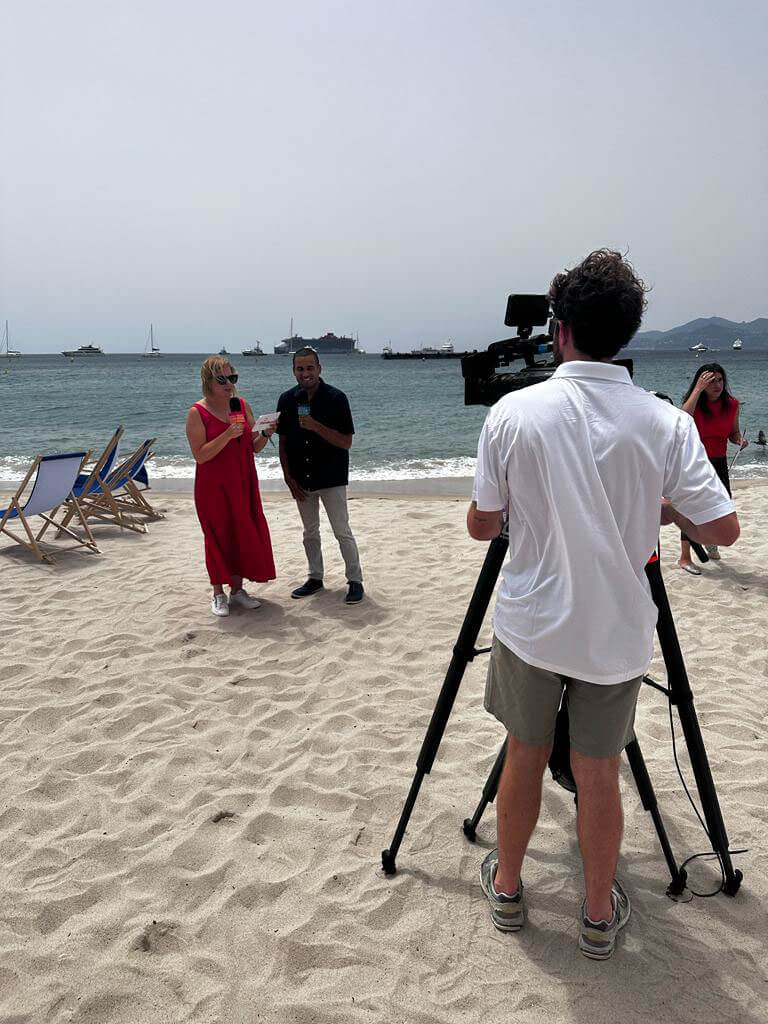 A woman in a red dress and a man being filmed during an interview on a sunny beach.