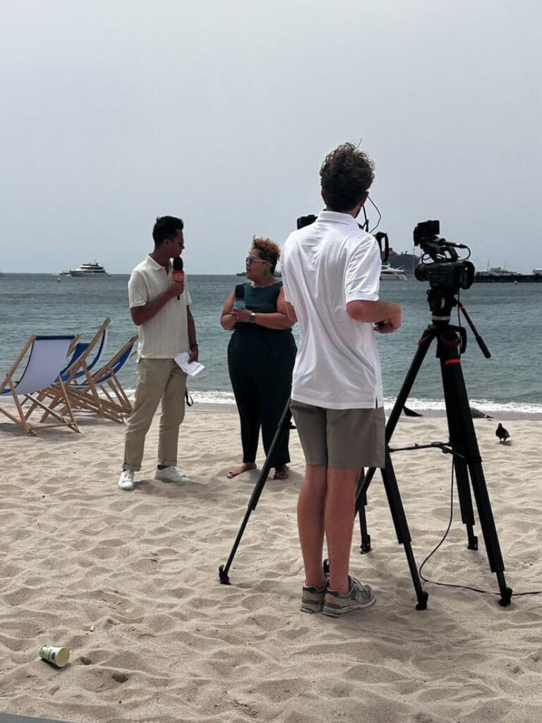 Two speakers engaged in conversation with a film crew capturing the moment, set against a backdrop of sand and sea.