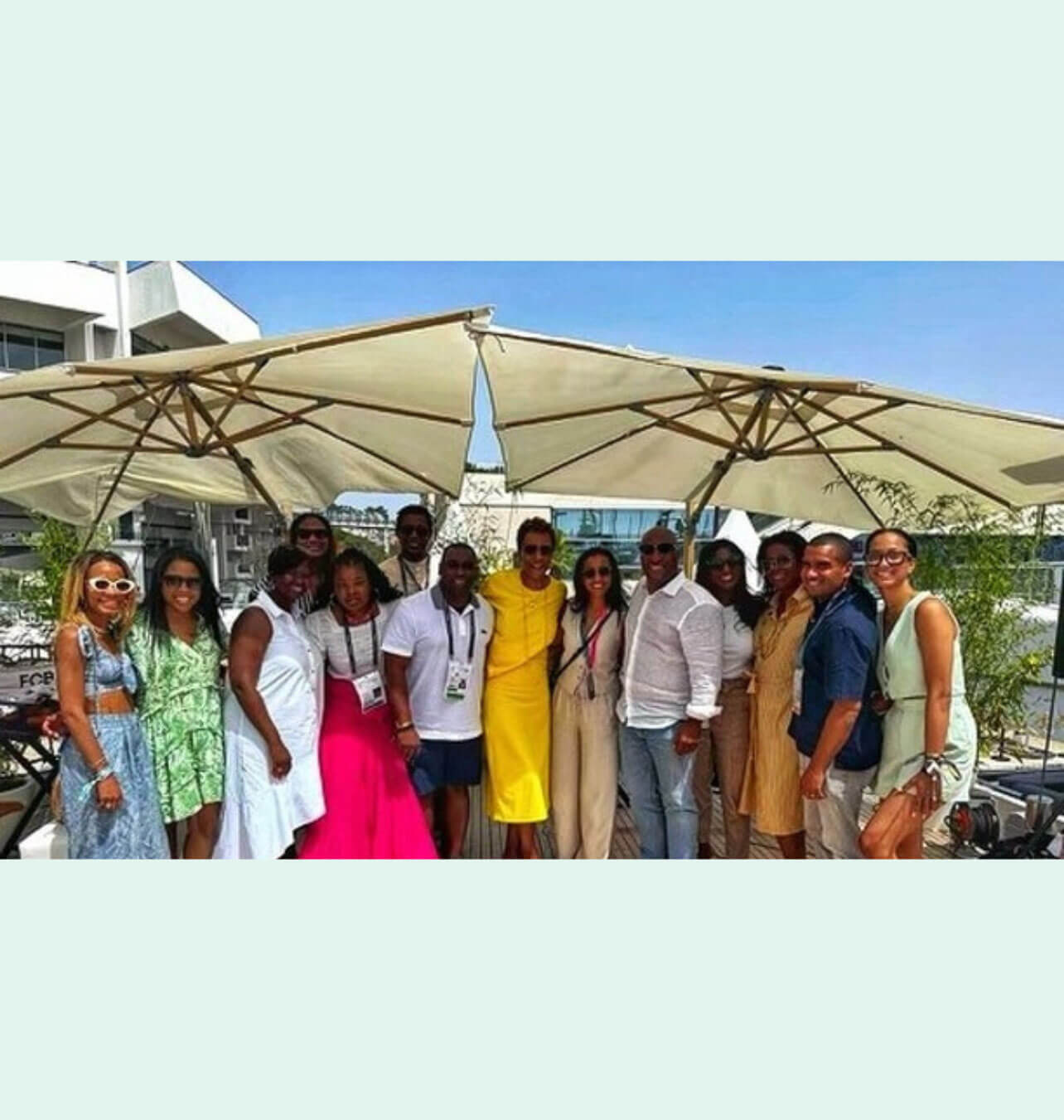 A large group of attendees posing together outdoors under patio umbrellas in a sunny setting.