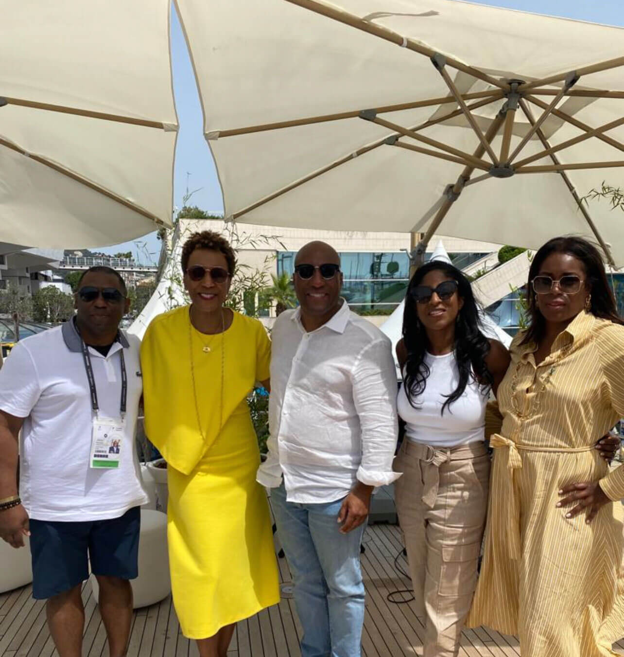 A group of five individuals posing together under patio umbrellas in a sunny outdoor setting.