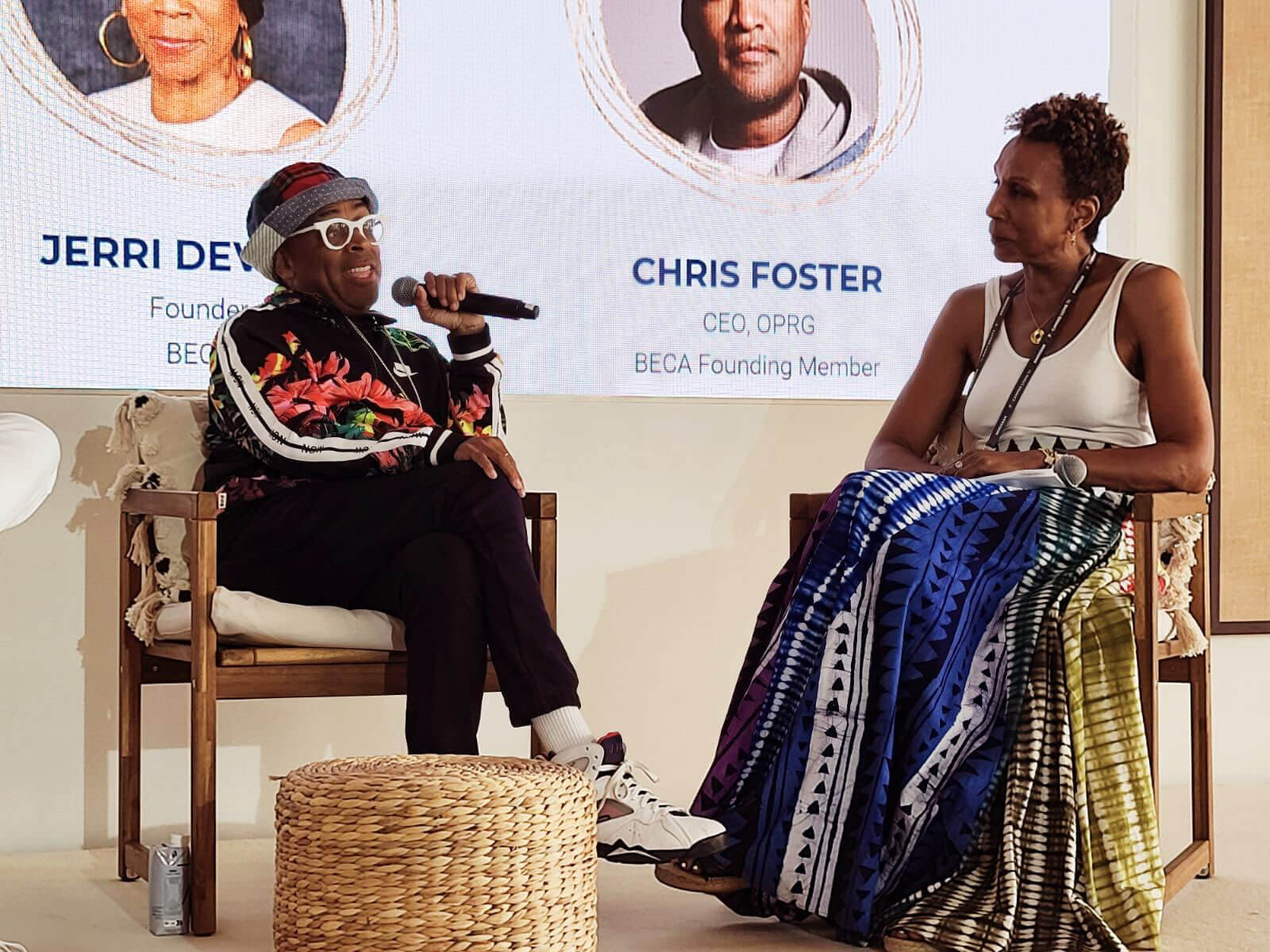 Two individuals seated on stage, one speaking into a microphone, with a screen in the background displaying names and titles.