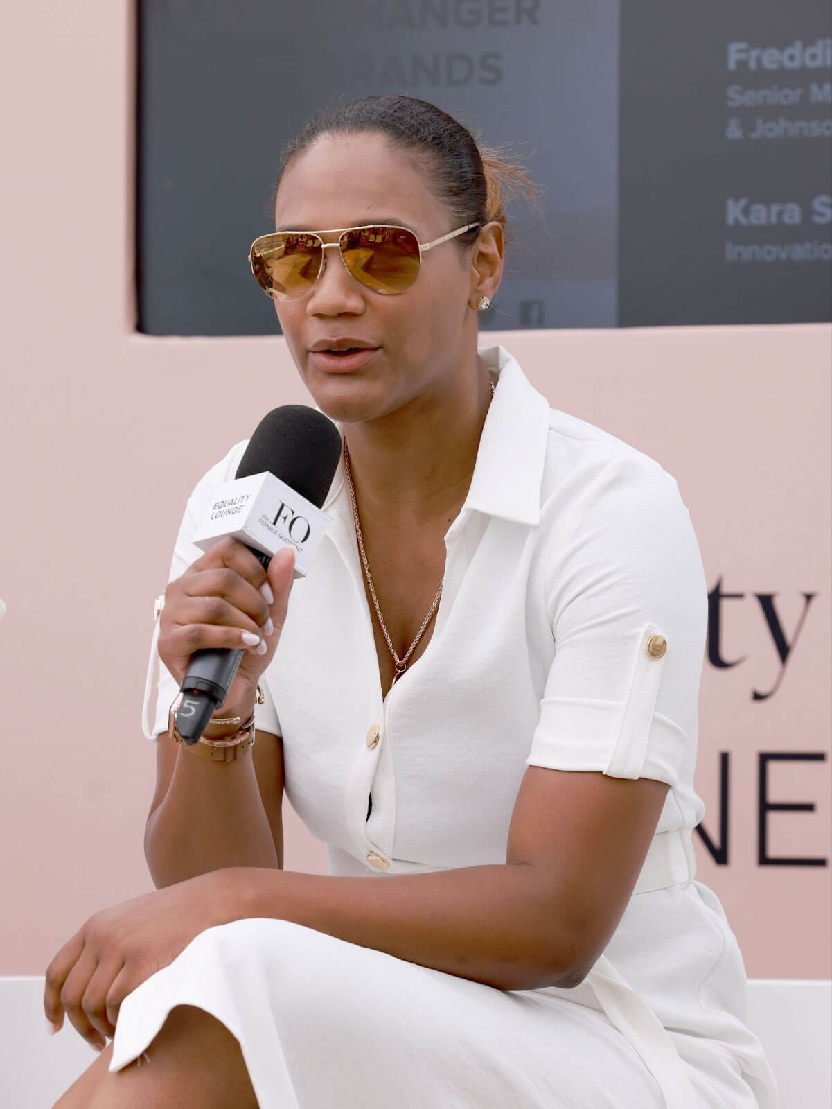 A woman in sunglasses and a white outfit speaking into a microphone with a backdrop displaying event details.