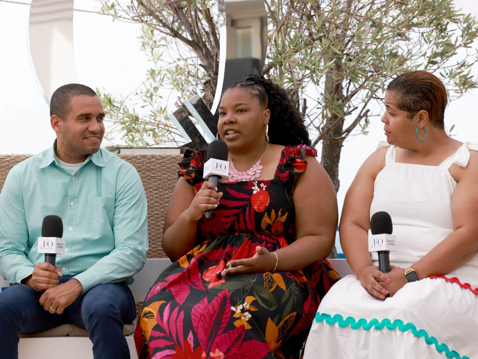 Three individuals seated on a stage holding microphones, participating in a discussion with a backdrop of greenery.