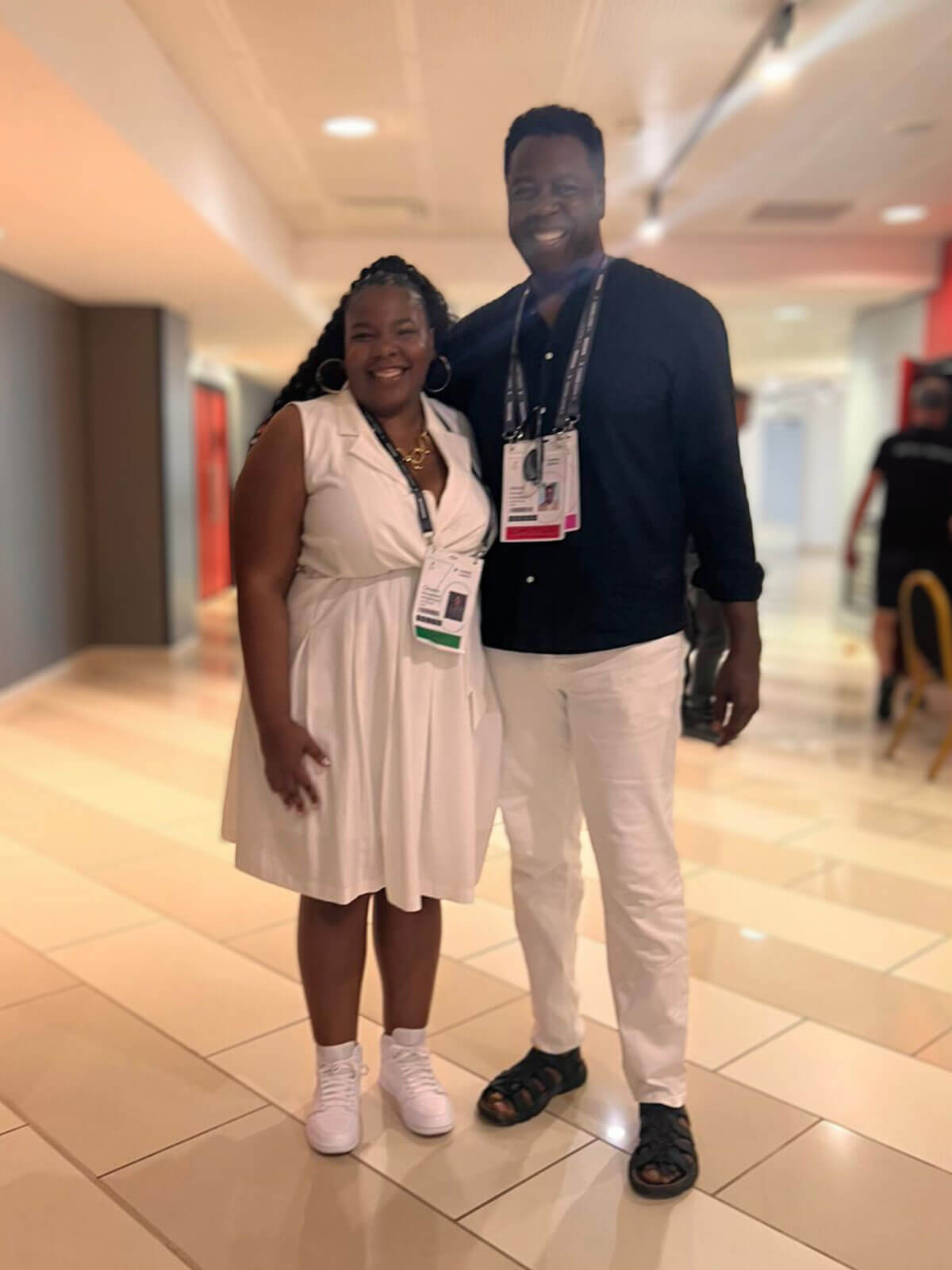 A man and a woman smiling and standing together in a brightly lit hallway, wearing casual yet stylish outfits.