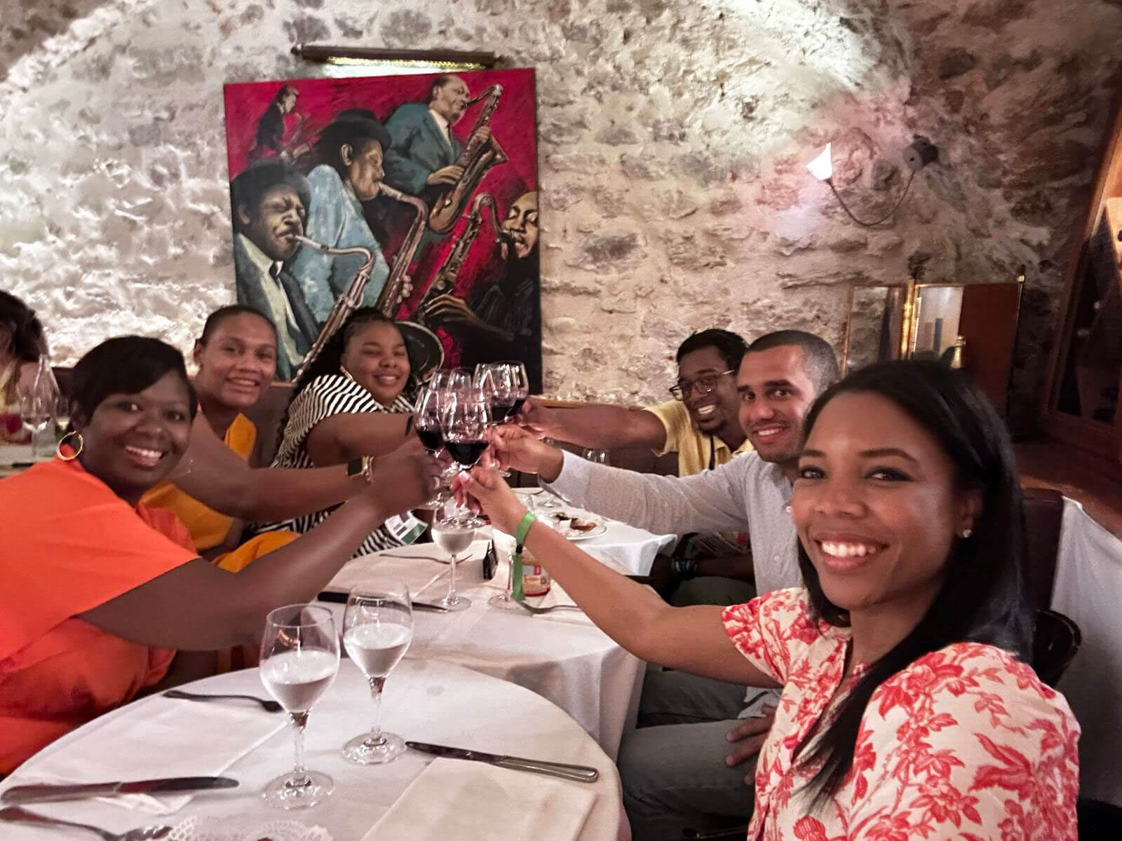 A group of people toasting with glasses of red wine at a dinner table in a warmly lit setting with artistic wall decor in the background.