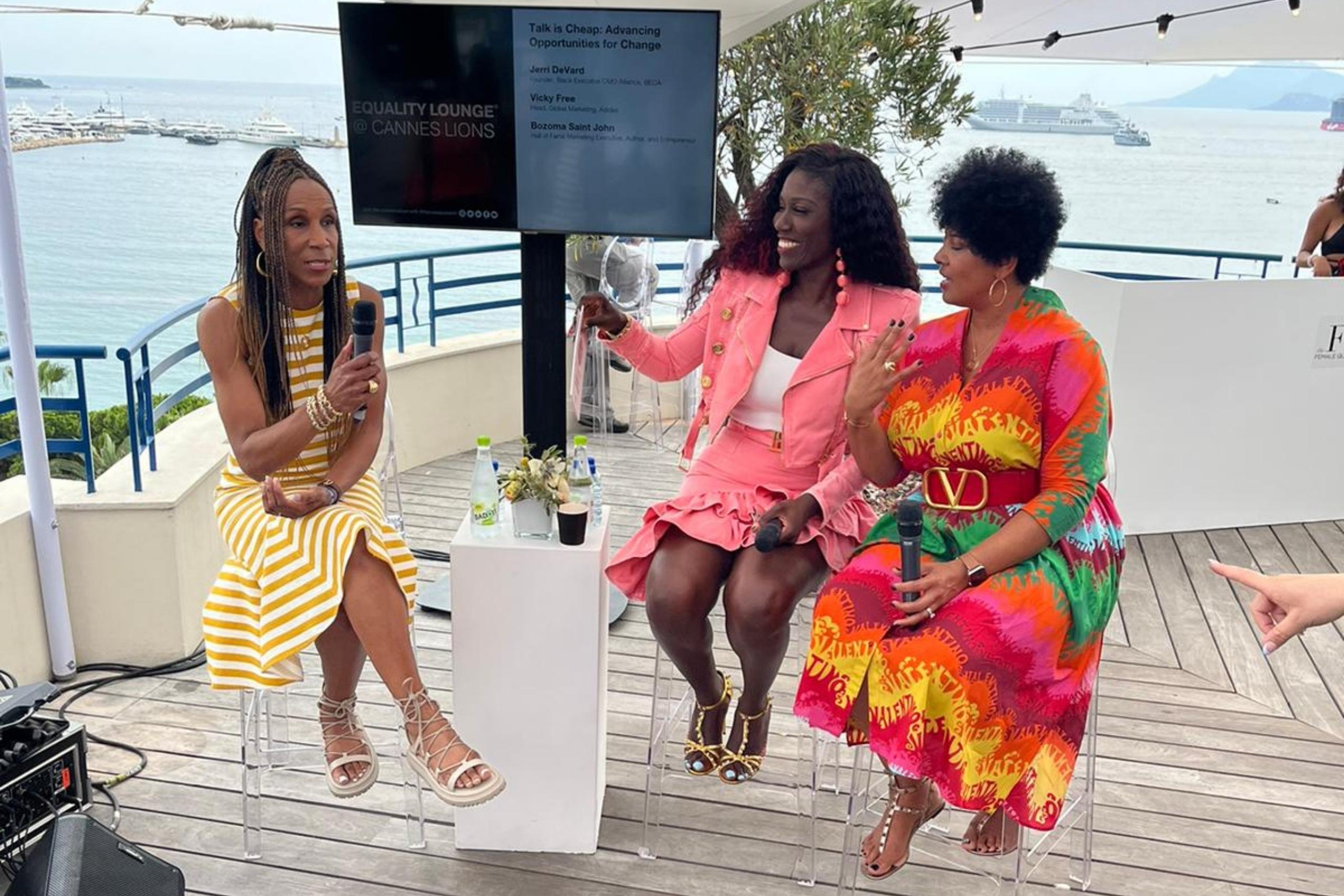 Three women engaged in a panel discussion at an outdoor event, with a marina and yachts in the background. Each is dressed in vibrant, stylish attire.