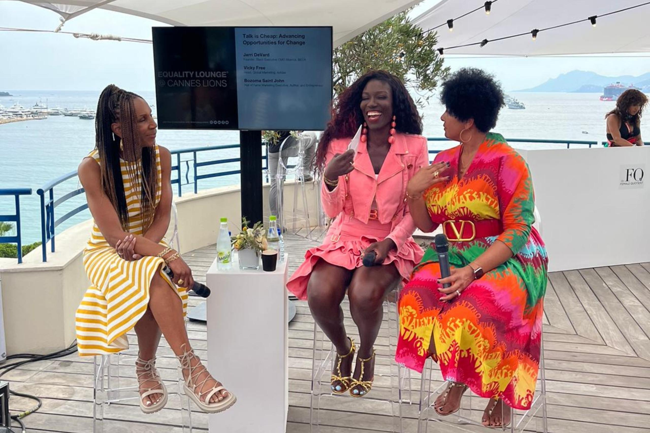 Three women seated on a panel at an outdoor event with a scenic ocean view. They are engaged in a lively discussion, holding microphones and laughing.