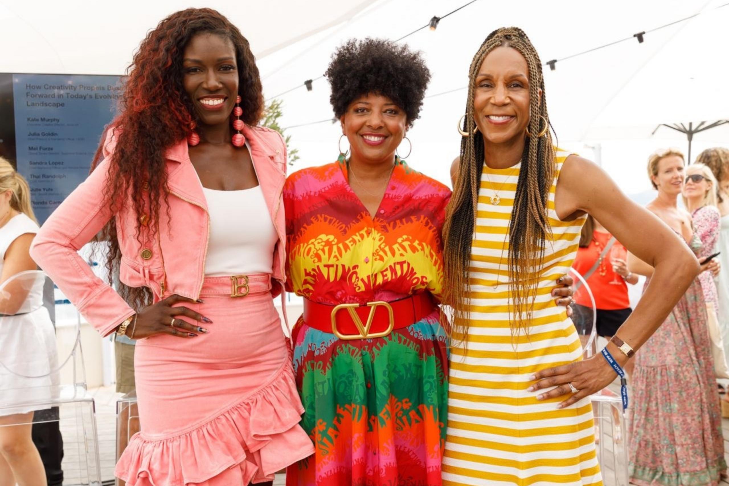 Three women standing side by side under an outdoor canopy, dressed in vibrant and stylish outfits, smiling warmly at the camera.