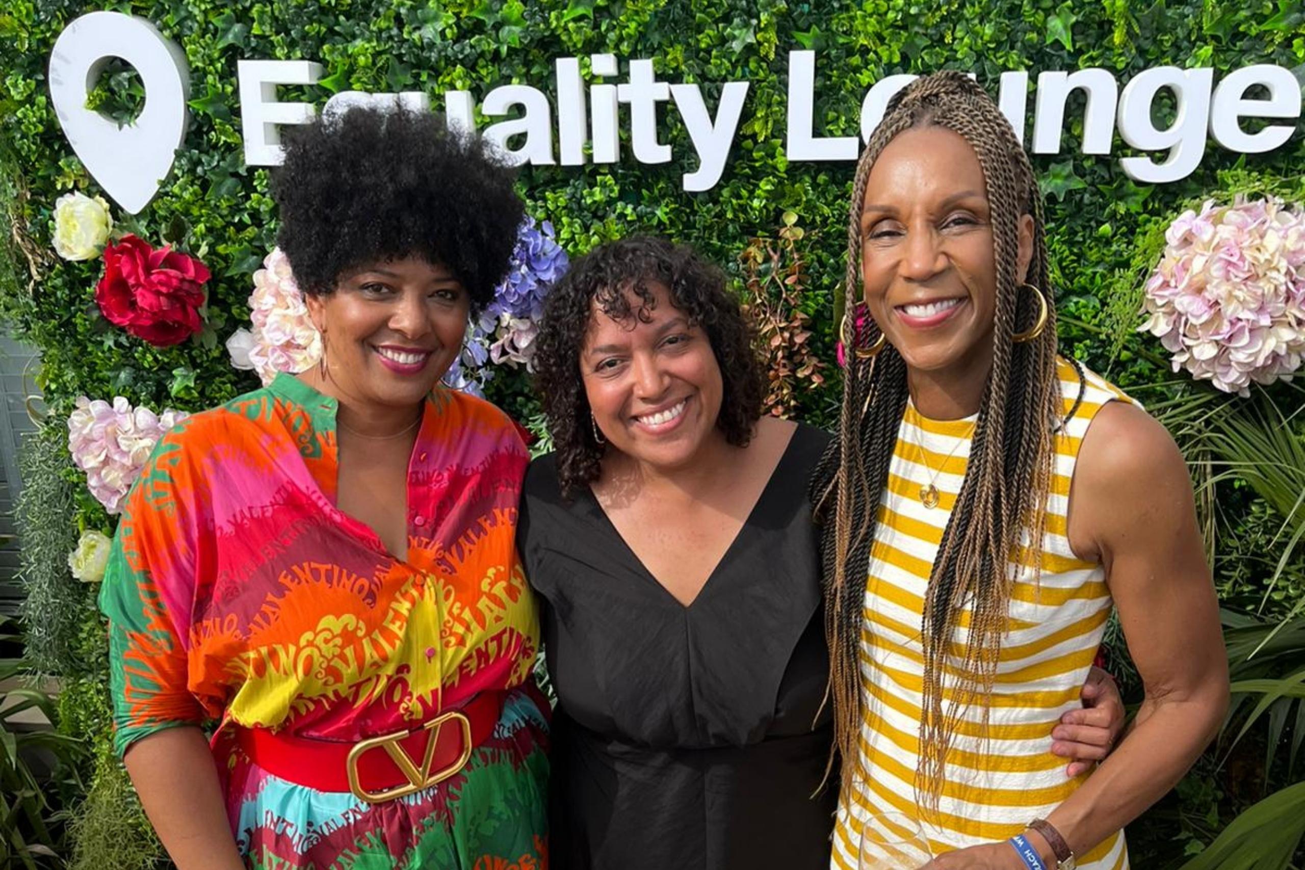 Three women smiling and posing together in front of a lush greenery wall adorned with colorful flowers and the text "Equality Lounge."