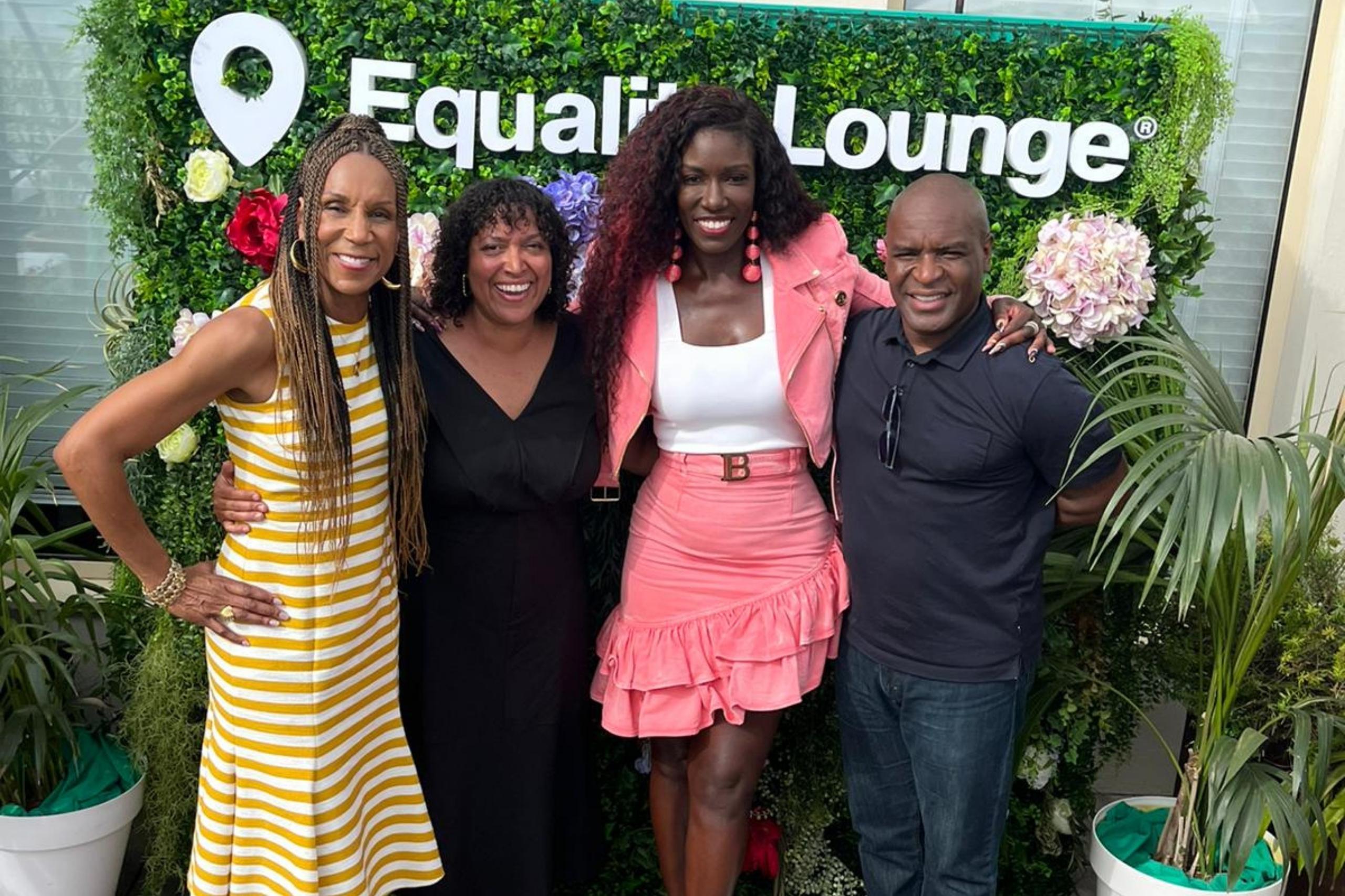A group of four people standing together, smiling in front of a greenery backdrop with the text "Equality Lounge." The group includes two women on the left, one wearing a yellow striped dress and the other in a black outfit, alongside a woman in a pink outfit and a man in a black shirt.
