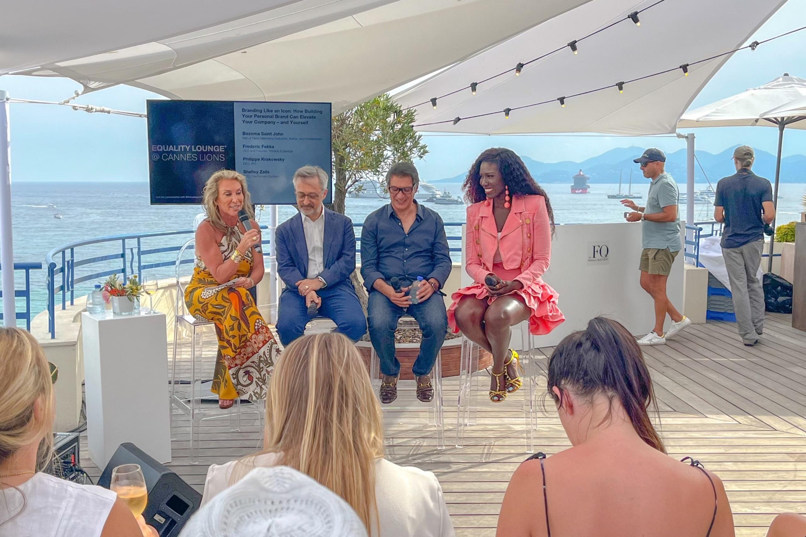 A panel discussion taking place outdoors by the water. Four panelists are seated on clear chairs, with an audience in the foreground. The background includes a screen displaying event details and scenic ocean views.