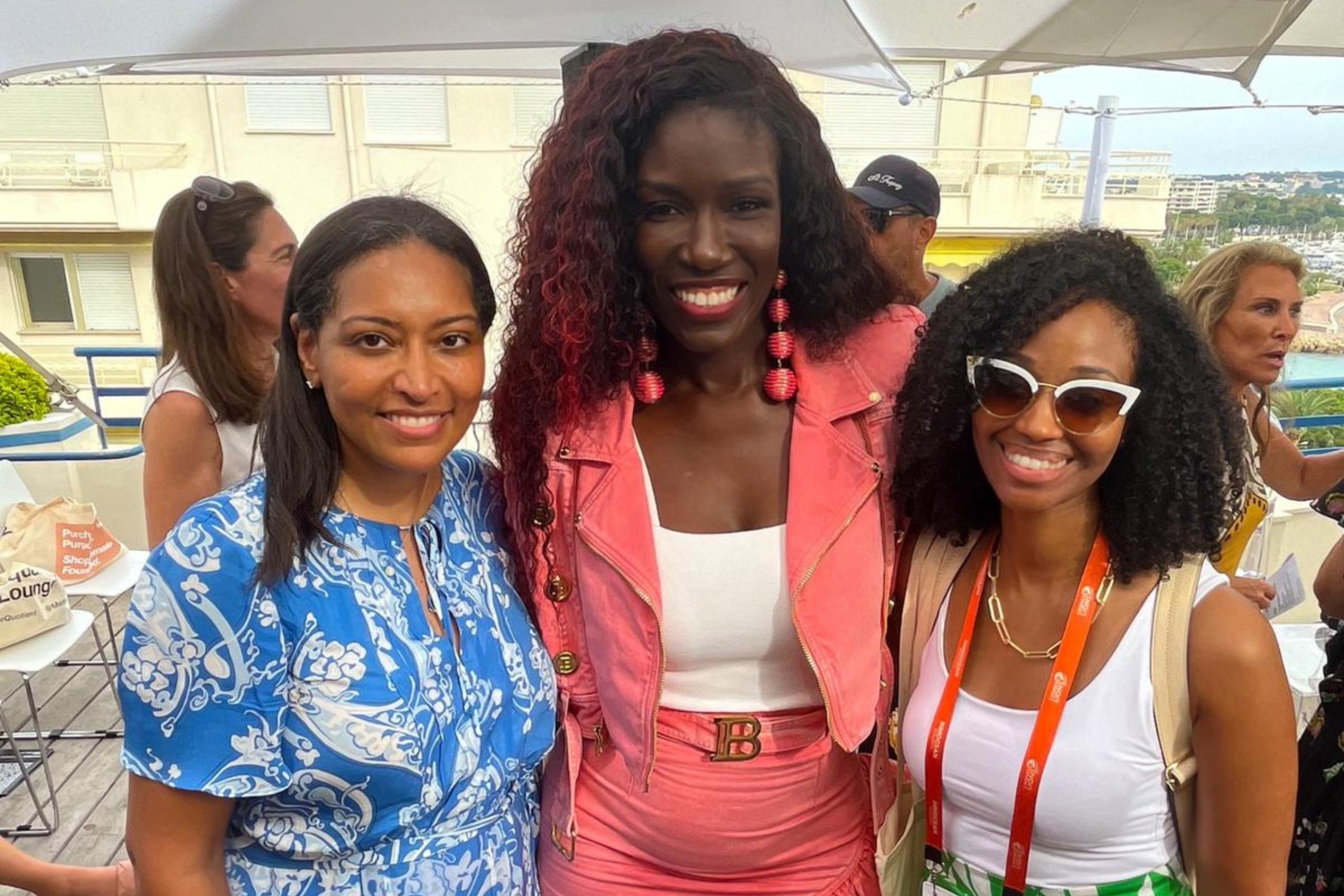 A woman in a pink outfit stands between two women, one wearing a blue patterned dress and the other in white and green attire, all smiling together at an outdoor event.
