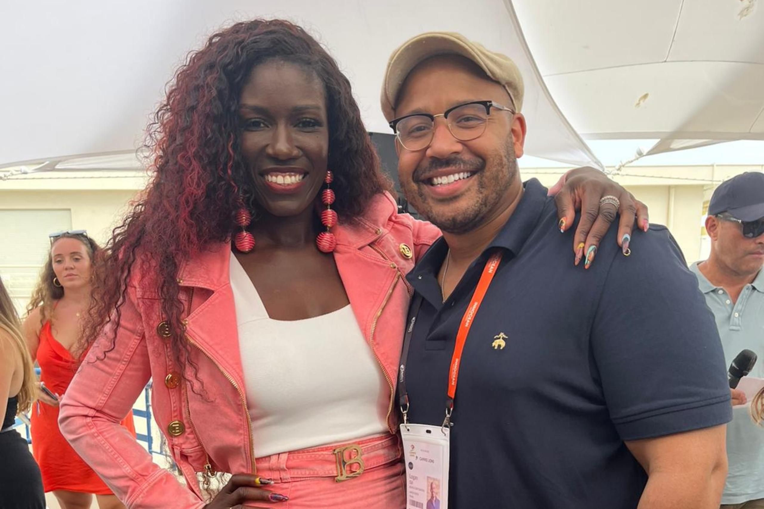 A man in a navy blue polo and beige cap poses with a smiling woman wearing a pink jacket and matching earrings. They are outdoors with other people in the background.