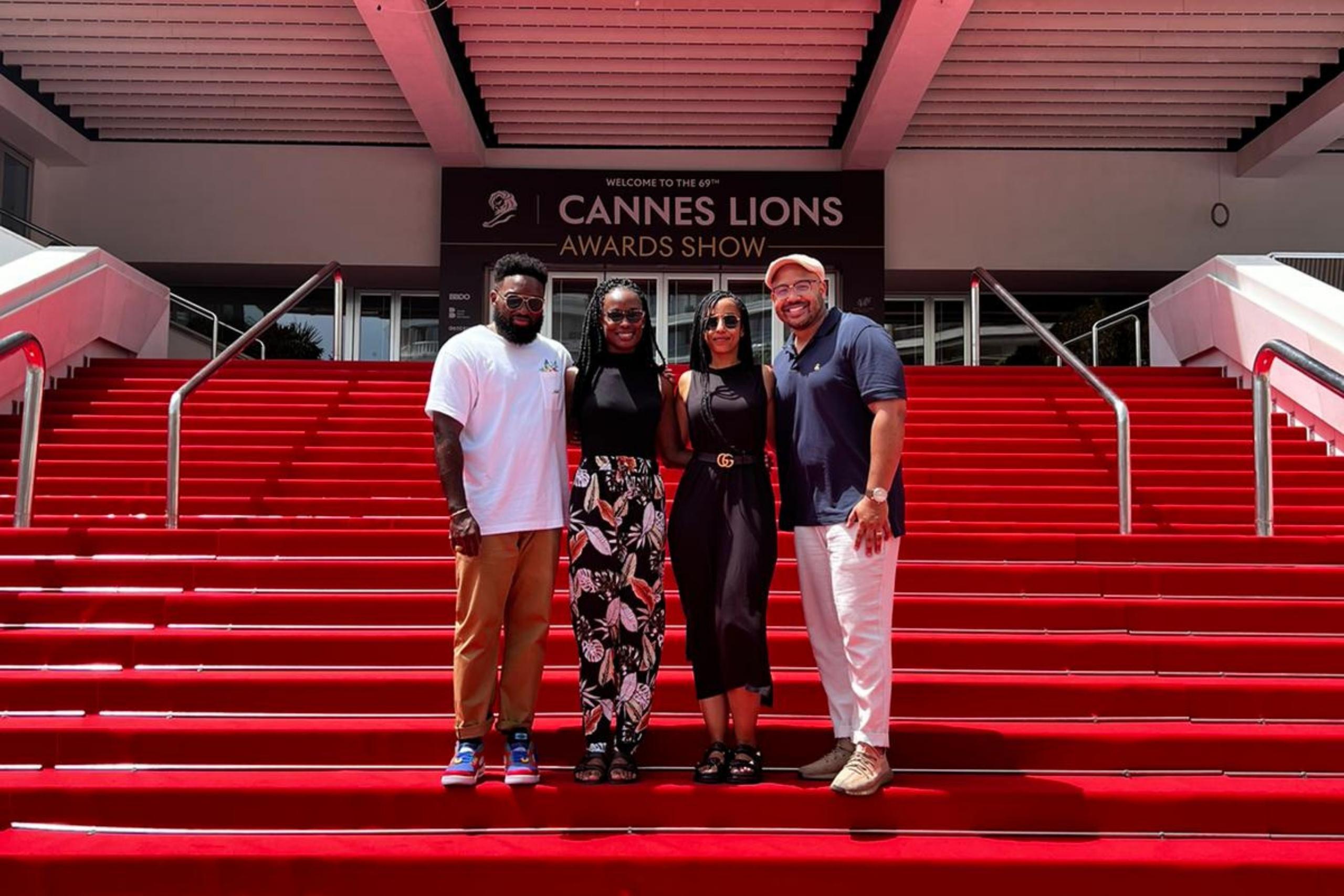 Four individuals pose together on red carpeted stairs in front of the "Cannes Lions Awards Show" sign. The group includes two men in casual attire and two women dressed in black and patterned outfits.