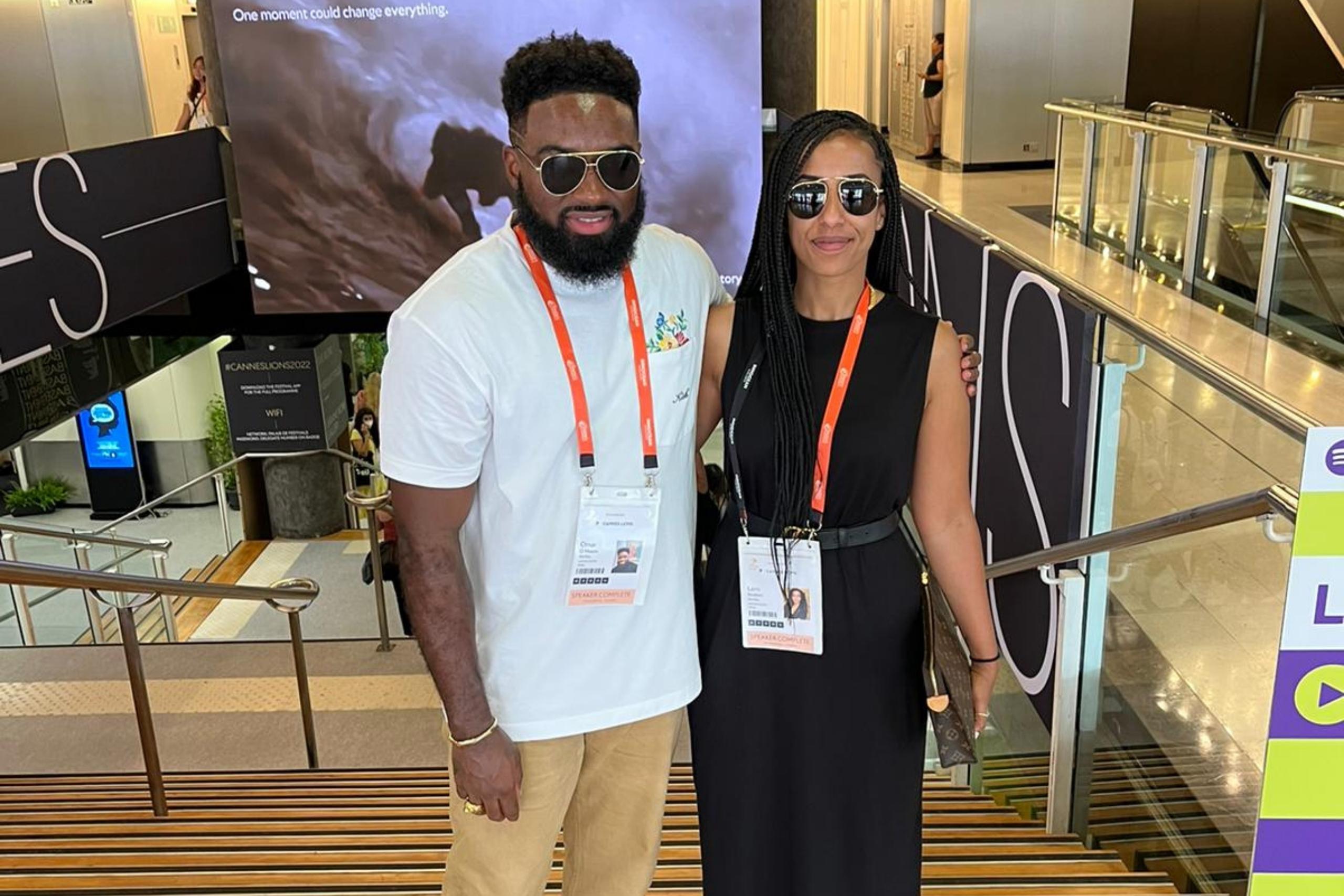 A man in a white shirt and beige pants stands beside a woman in a black dress. Both wear sunglasses and event badges, posing on a staircase with a large screen in the background.