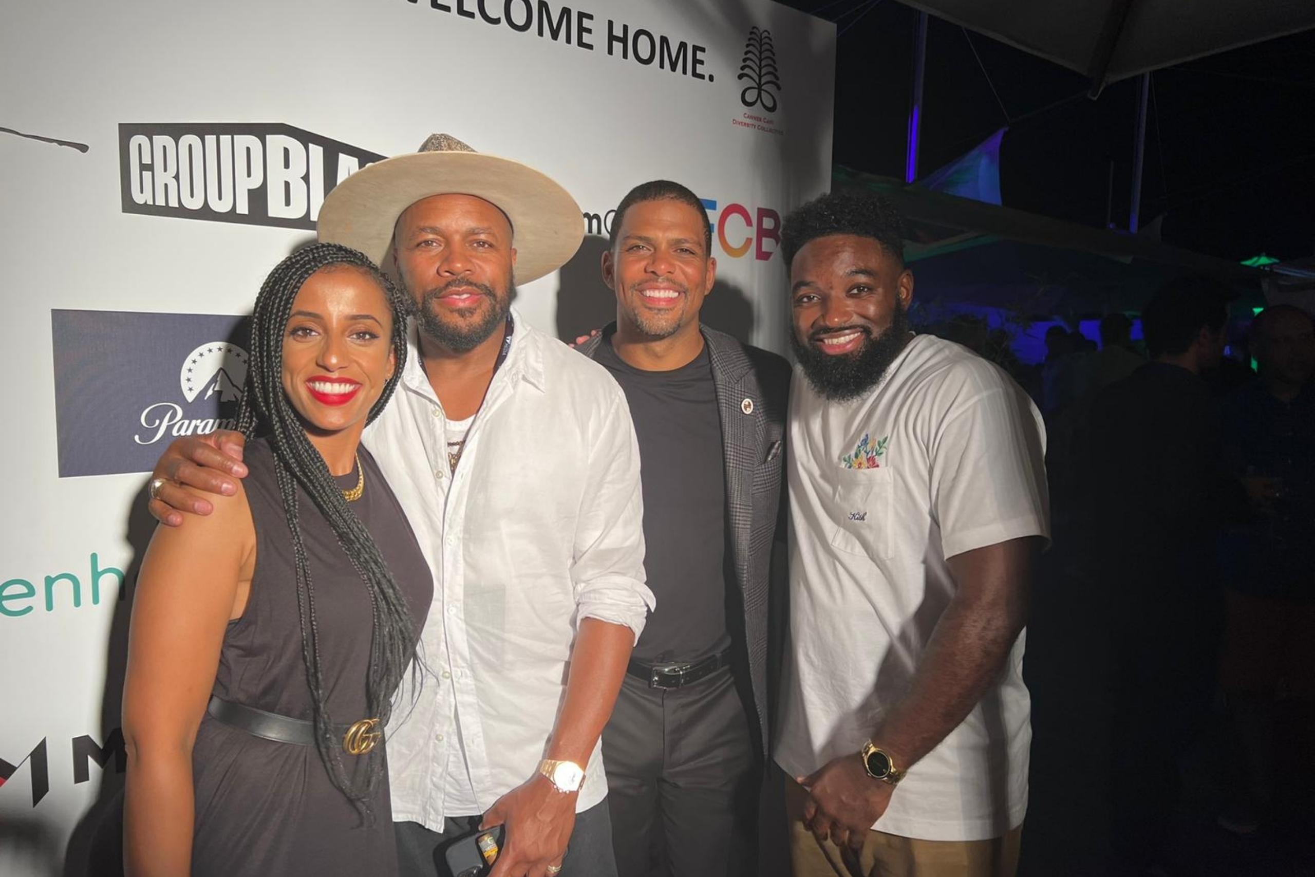 Four individuals pose together at an event with a branded backdrop. The group includes two men, one in a white shirt and hat, and two women, one wearing a black dress and braids.