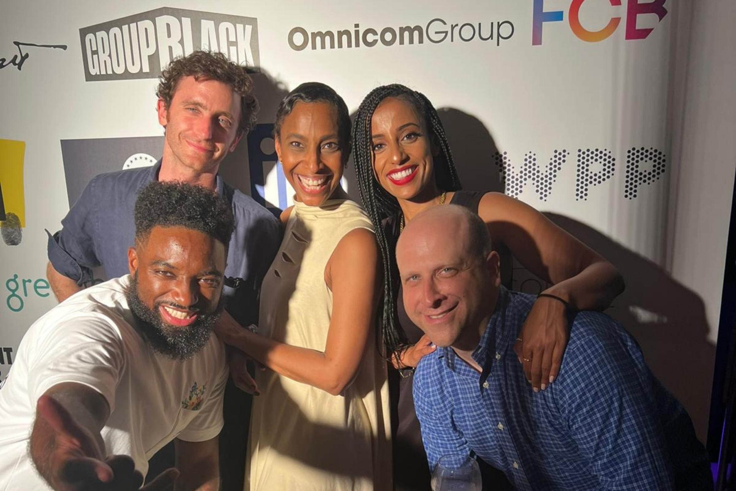 A group of five people posing together against a branded backdrop with logos like "Group Black" and "Omnicom Group." They are smiling, with one man leaning forward playfully.