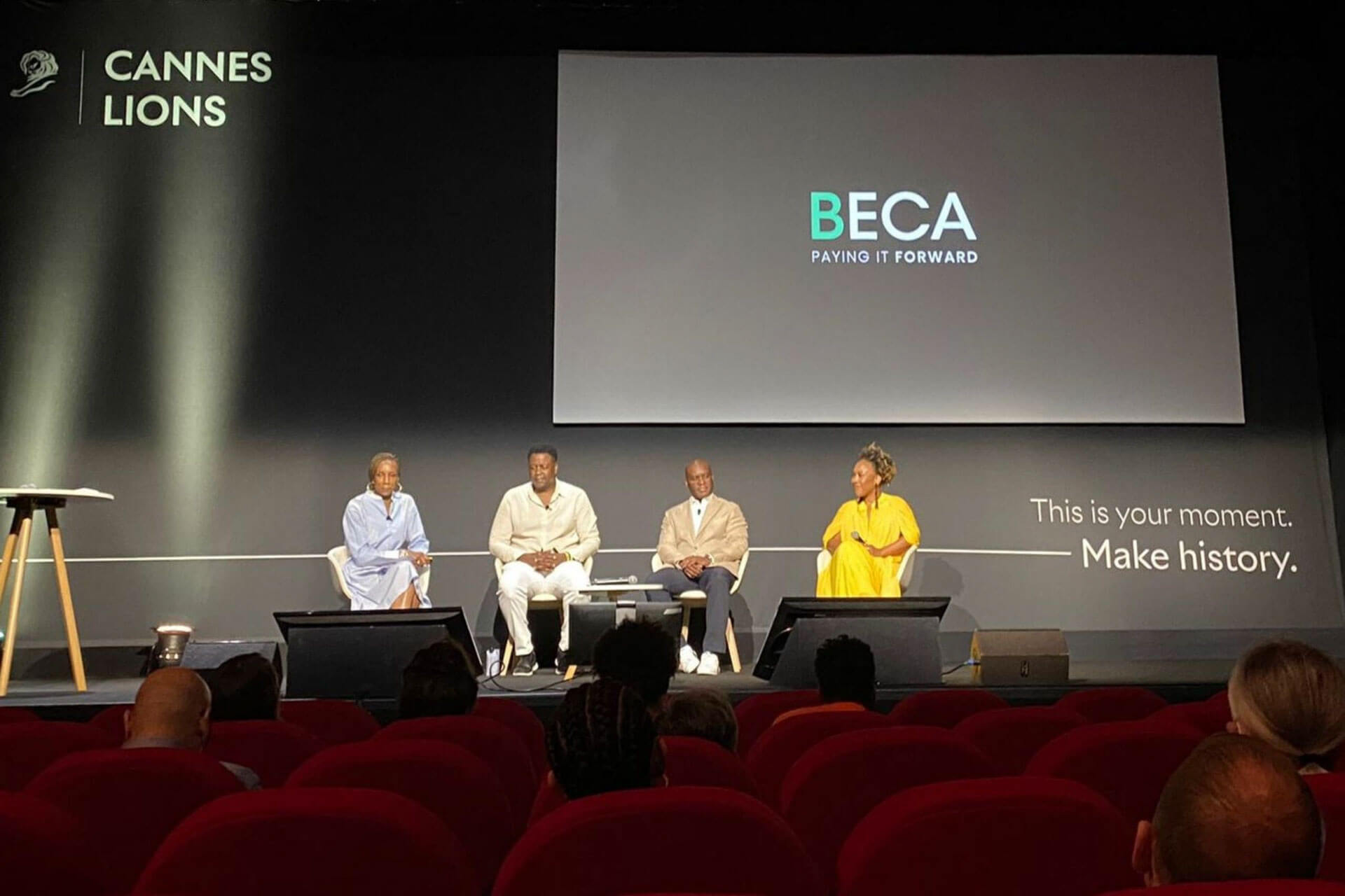 A panel of four individuals seated on a stage at the Cannes Lions event, with "BECA Paying It Forward" displayed on the screen behind them. The audience watches from red theater seating.