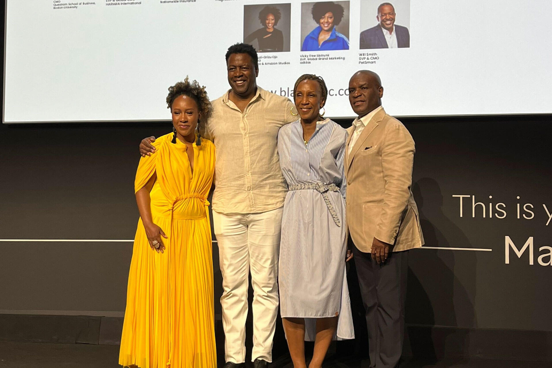 A close-up group photo of four individuals on a stage, smiling warmly. The group includes two women, one in a yellow dress and one in a striped blue outfit, and two men in beige suits.