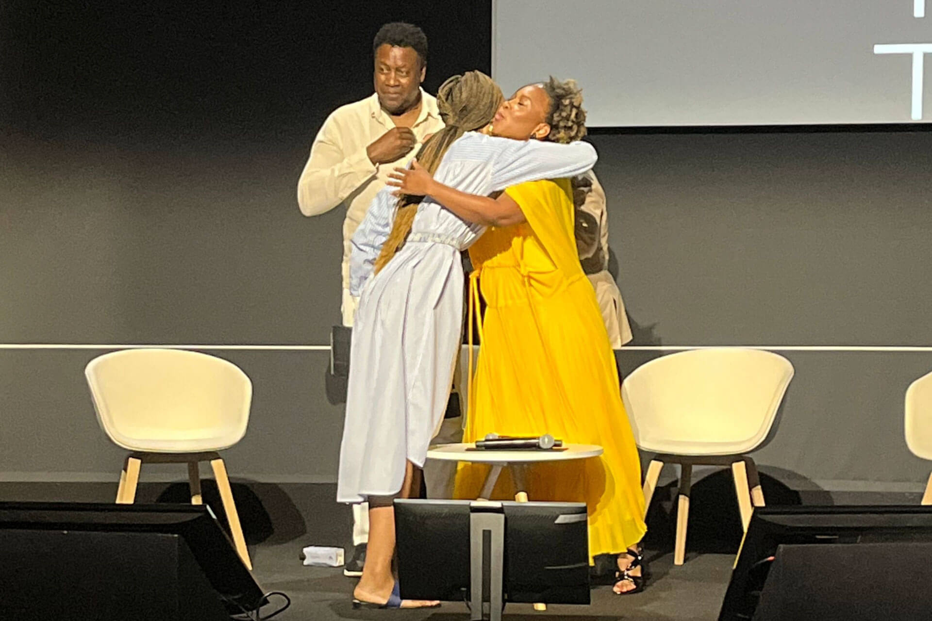 Three individuals on a stage; two women embrace warmly, one wearing a yellow dress, the other in a light blue outfit. A man stands behind them, smiling, with chairs and a small table in the background.