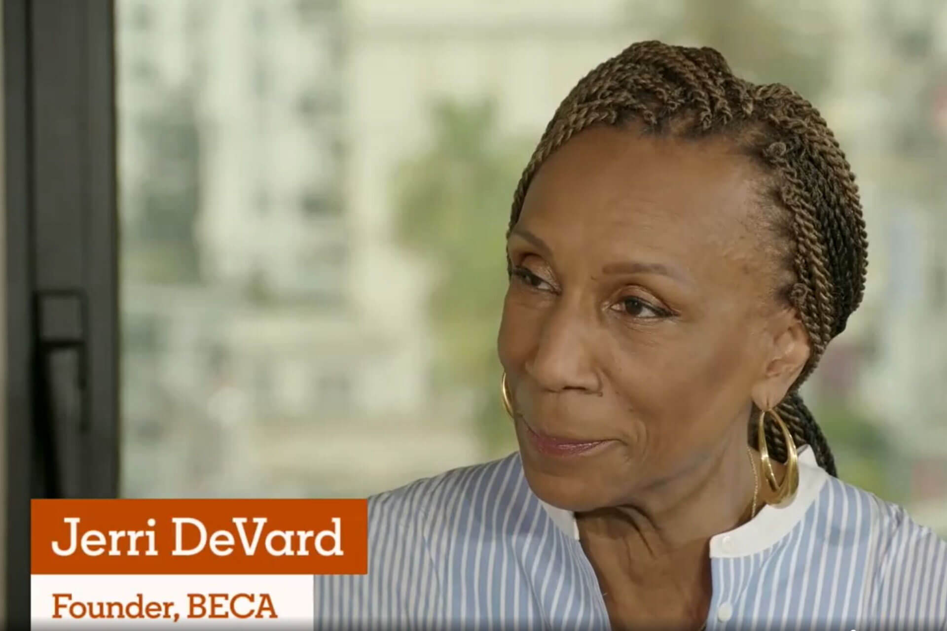 A close-up of a woman with braided hair and gold hoop earrings, identified as Jerri DeVard, founder of BECA, in an interview setting with a blurred urban background.