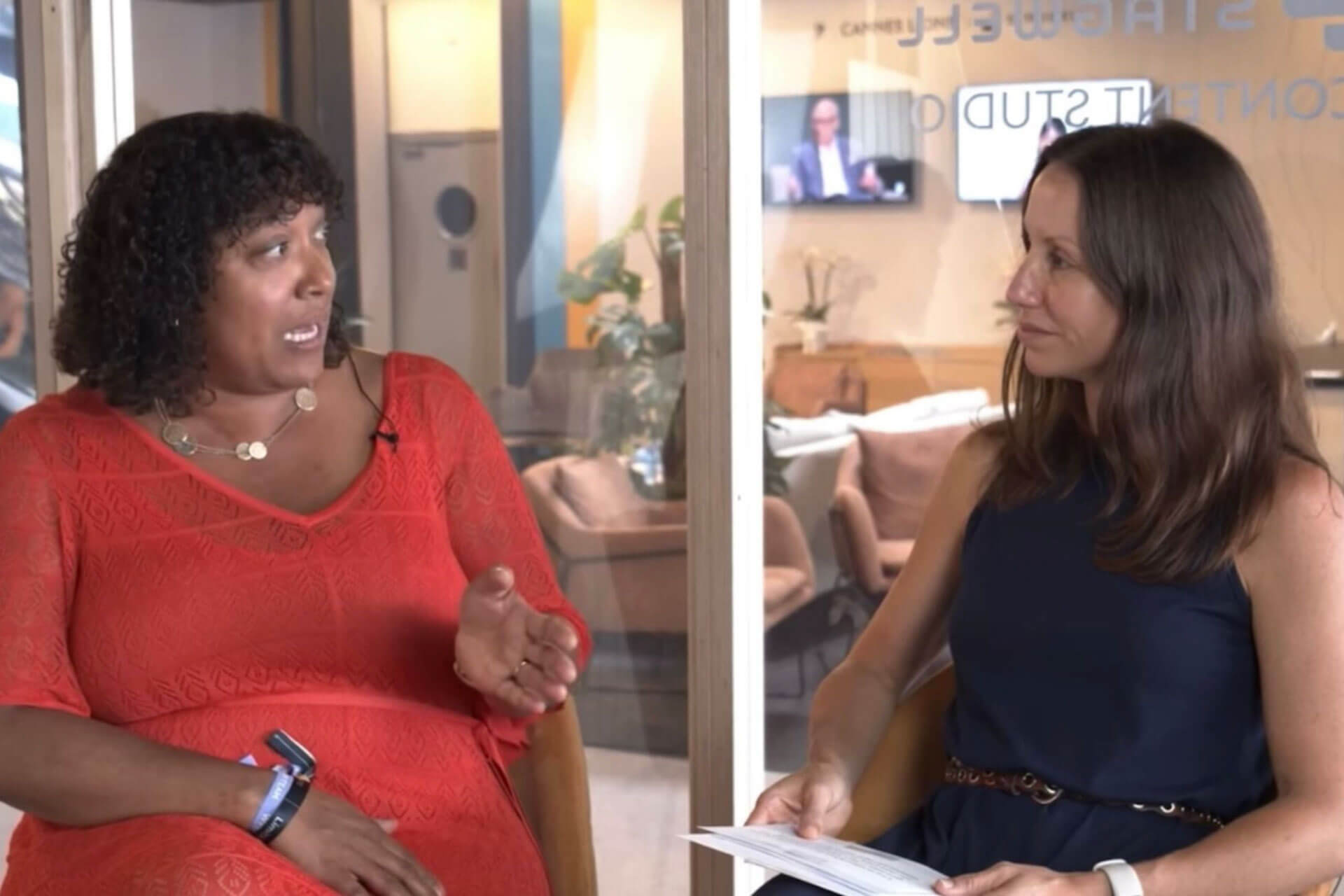 A woman in a red dress and a woman in a navy-blue dress sit across from each other in a studio-style setting, engaged in an animated discussion.