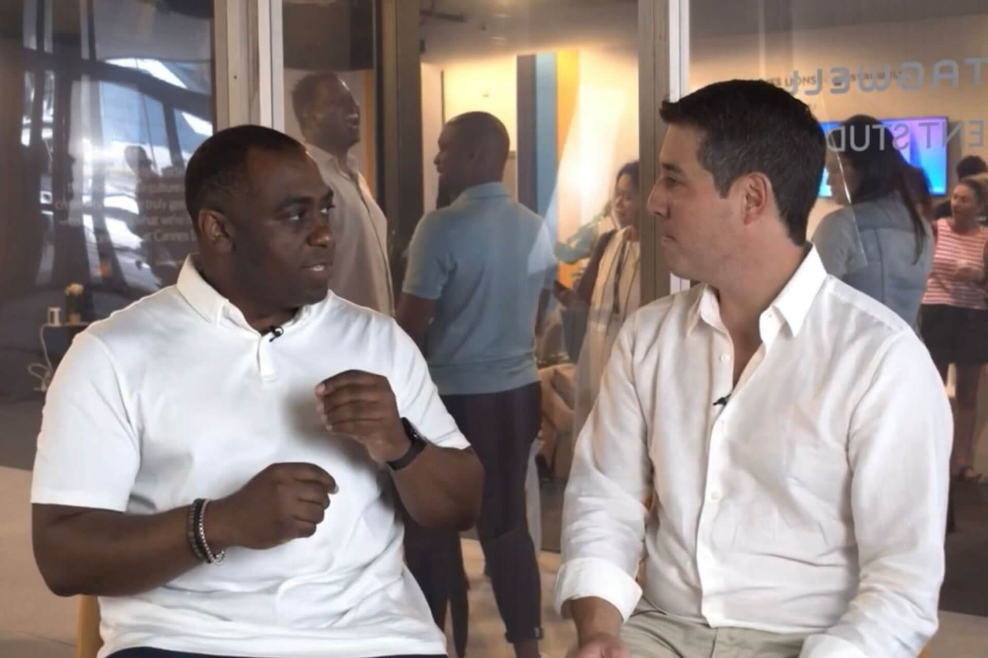 Two men in white shirts converse energetically in a modern indoor setting with glass walls and people visible in the background.
