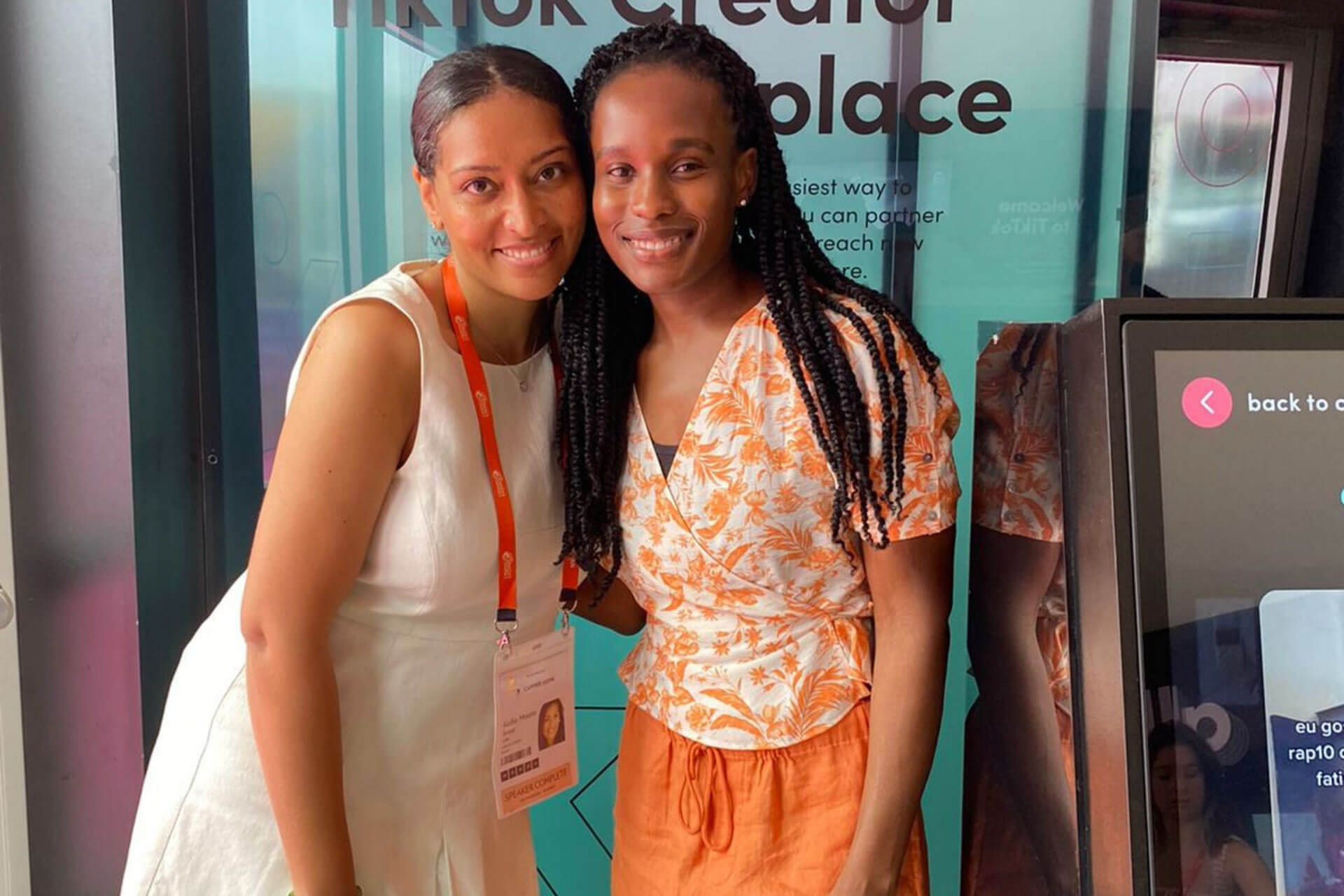 Two women, one in a white dress and the other in an orange floral top, stand closely together and smile in front of a vibrant TikTok Creator Marketplace banner.
