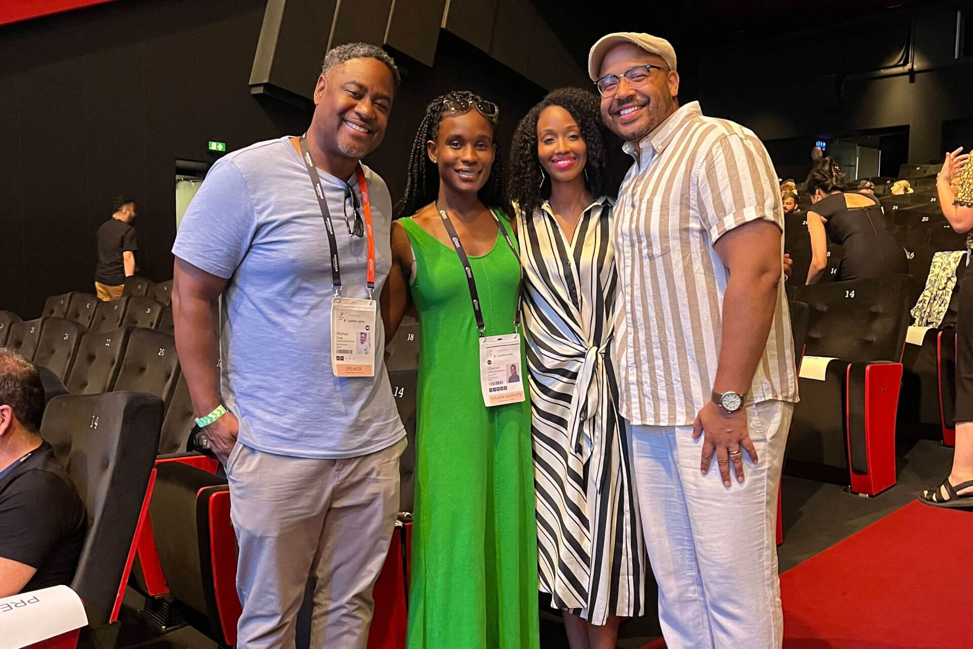 A group of four individuals in a theater, smiling for the camera, with event badges displayed on their lanyards.
