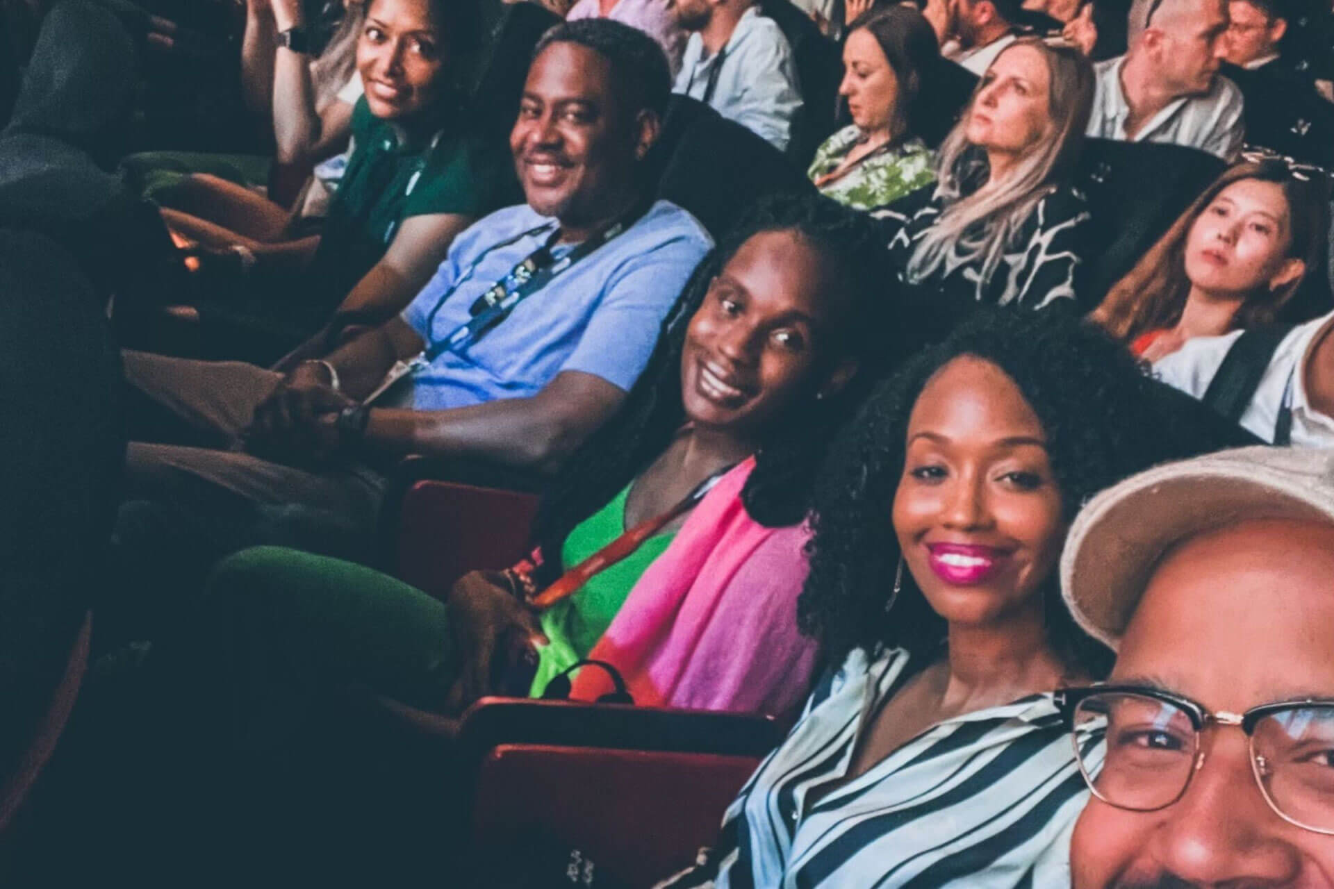 A row of people seated in a theater, smiling and enjoying the event, with others in the background.