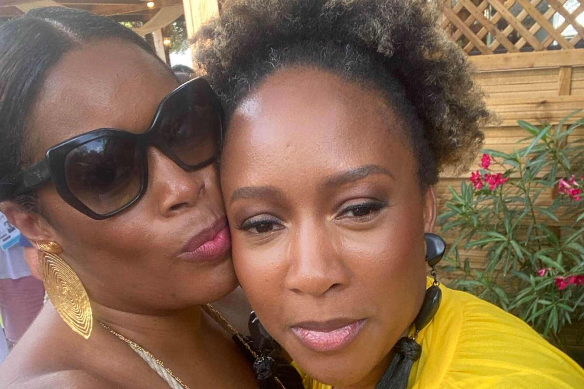 A close-up selfie of two women outdoors, one wearing large sunglasses and the other in a yellow top, both smiling warmly.