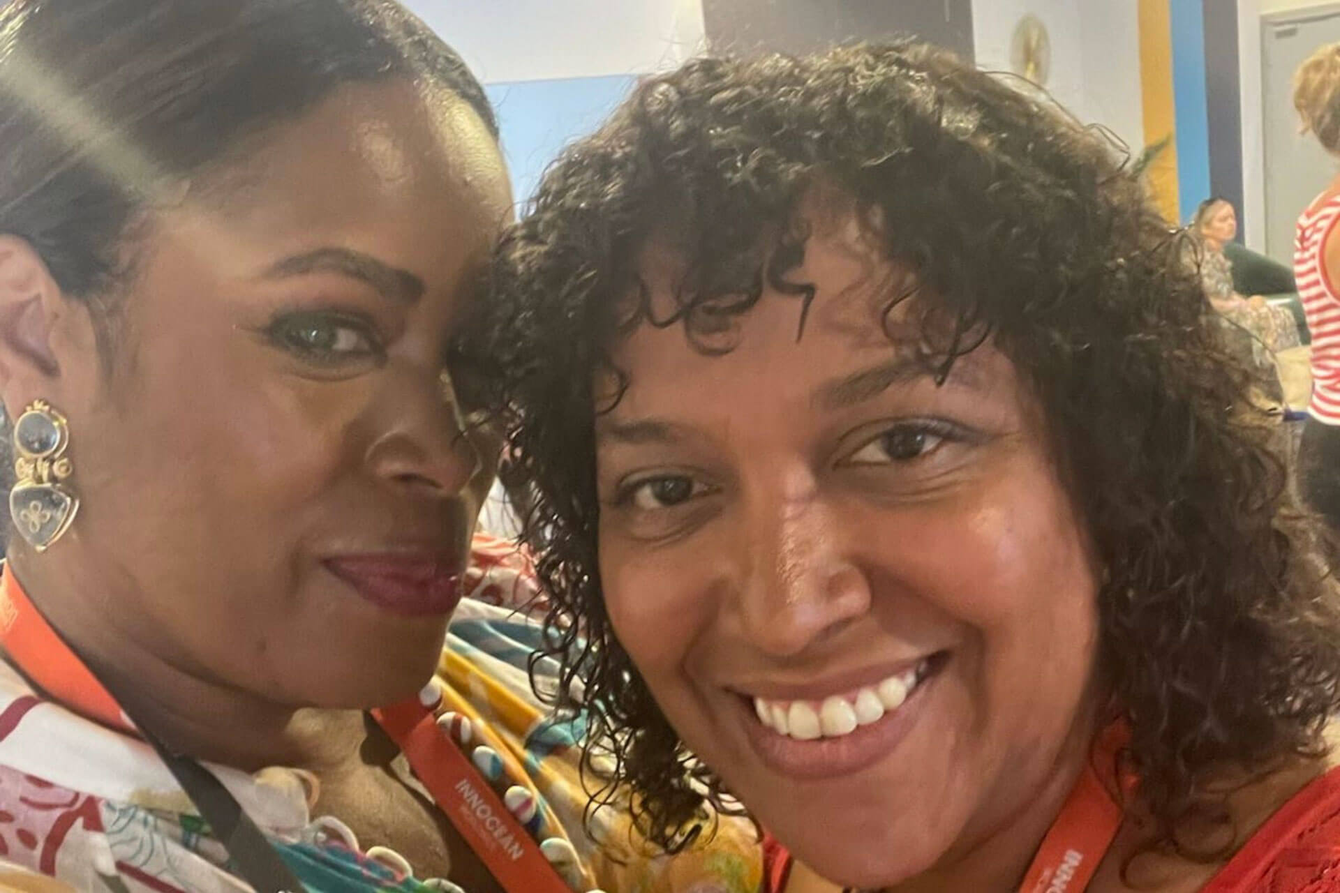 A close-up selfie of two women indoors, both smiling and wearing lanyards, with others in the background.