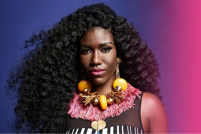 A portrait of a woman with voluminous curly hair and bold, colorful statement jewelry against a purple backdrop.