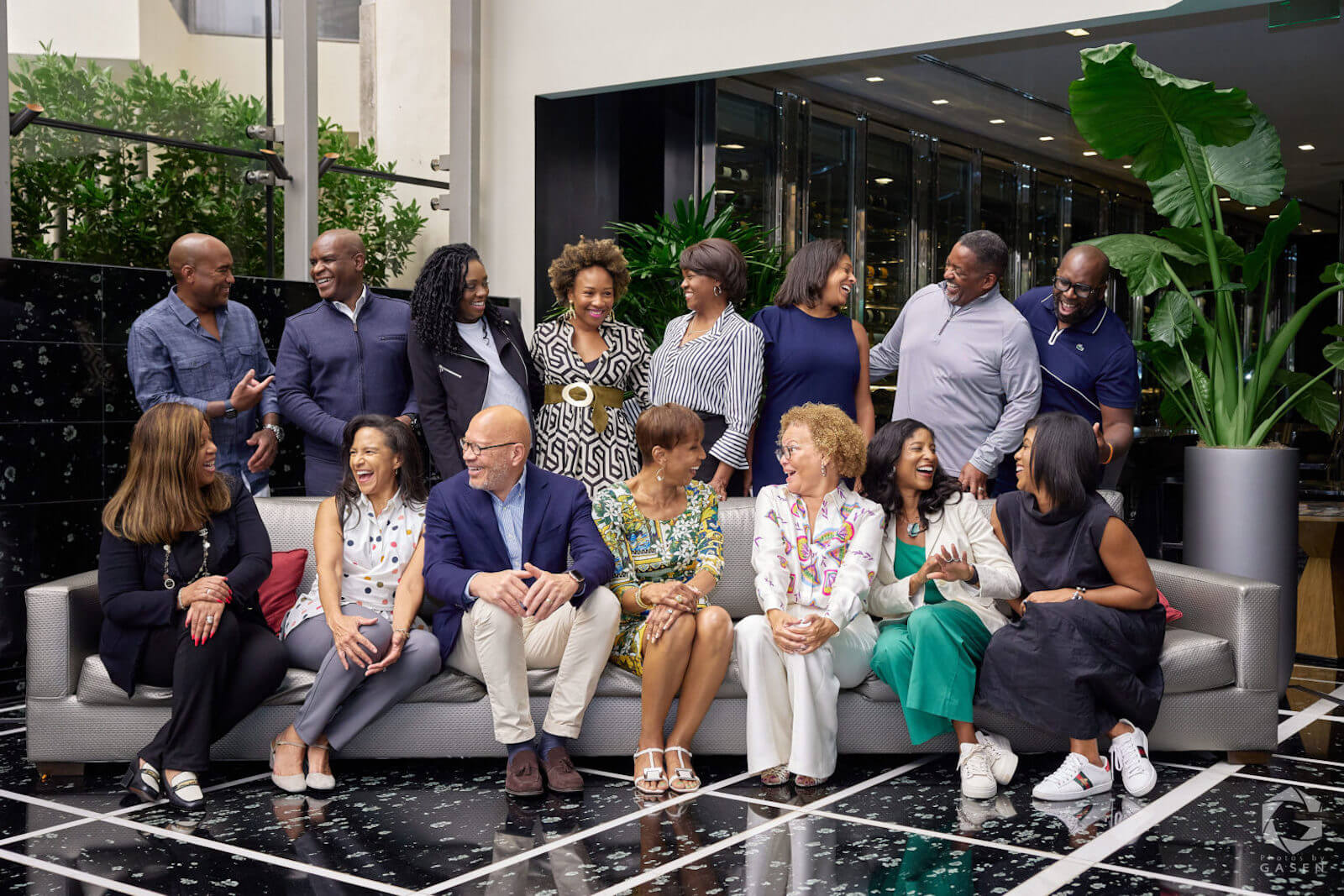 A well-dressed group of professionals laughing and conversing while seated and standing in a modern indoor space with greenery.