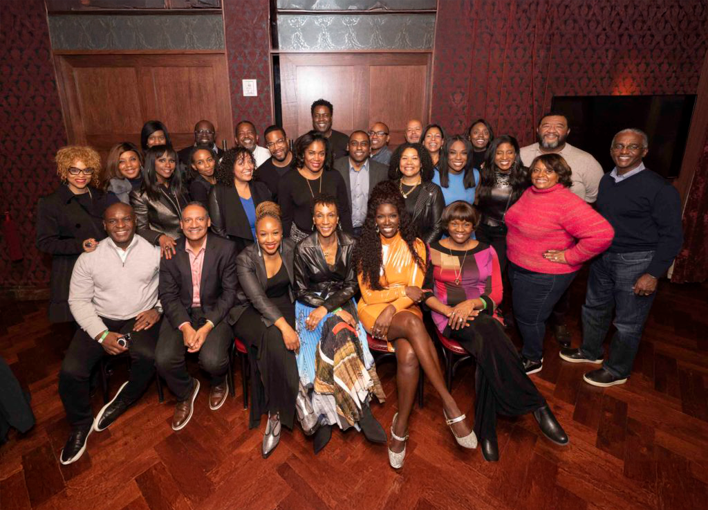 A large group of adults, some seated and some standing, smiles at the camera in a warm, wood-paneled room. They are dressed in a mix of business and festive attire.